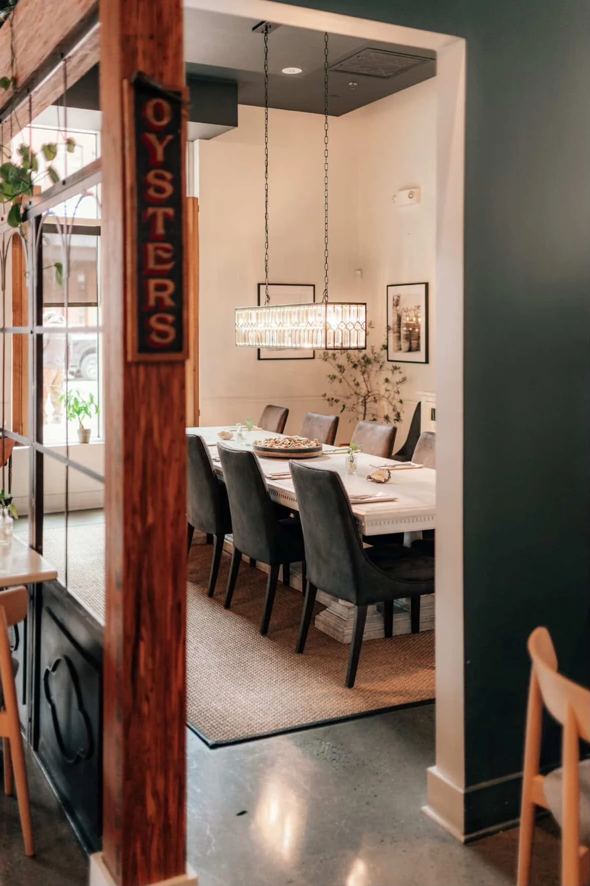 A dining room with a long white table, surrounded by black upholstered chairs, a modern chandelier, framed artwork on the walls, and a plant in the corner, viewed through a doorway with a wooden post labeled "OYSTERS."