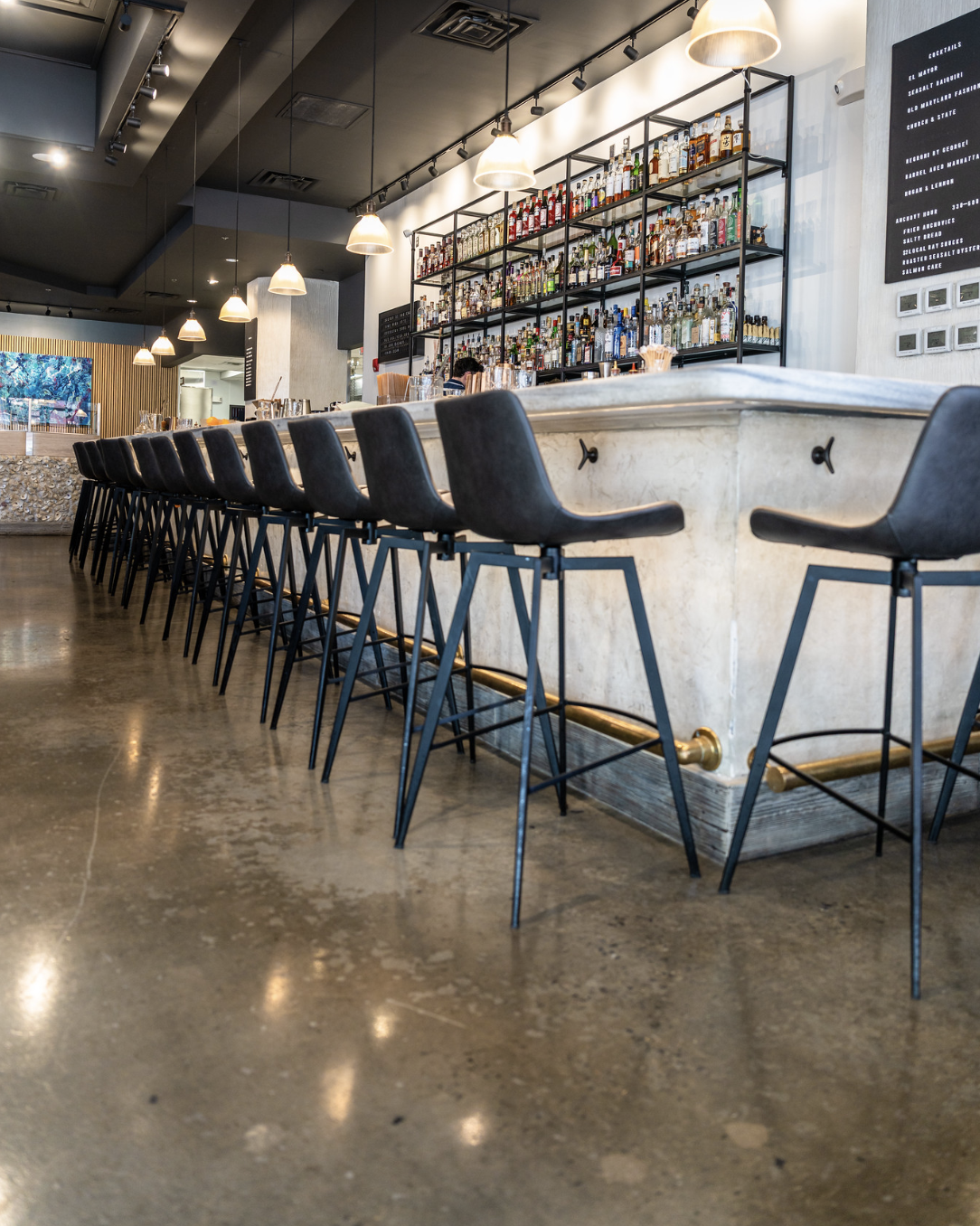 A modern bar with a white marble counter, black bar stools, and shelves filled with various bottles of alcohol behind the bar. Pendant lights hang from the ceiling, and the polished concrete floor reflects some light.