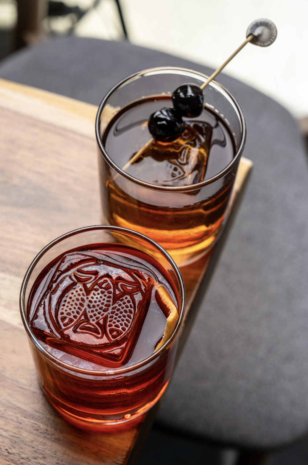 Two glasses of iced beverages with ice cubes; one has a lemon slice, and the other has a cherry and a cocktail stirrer with maraschino cherries.