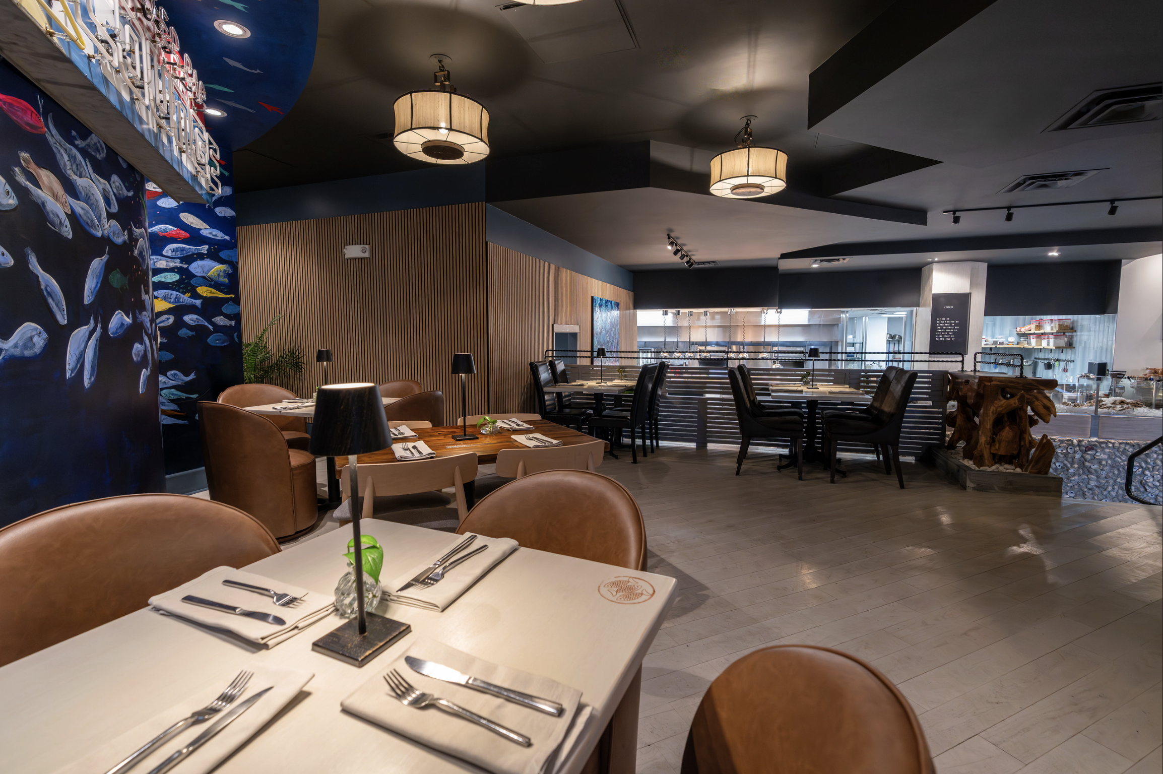 Interior view of a modern restaurant with wooden and dark-colored furniture, including tables set with silverware and small plants, decorative lighting fixtures on the ceiling, and a mural of fish on one wall.