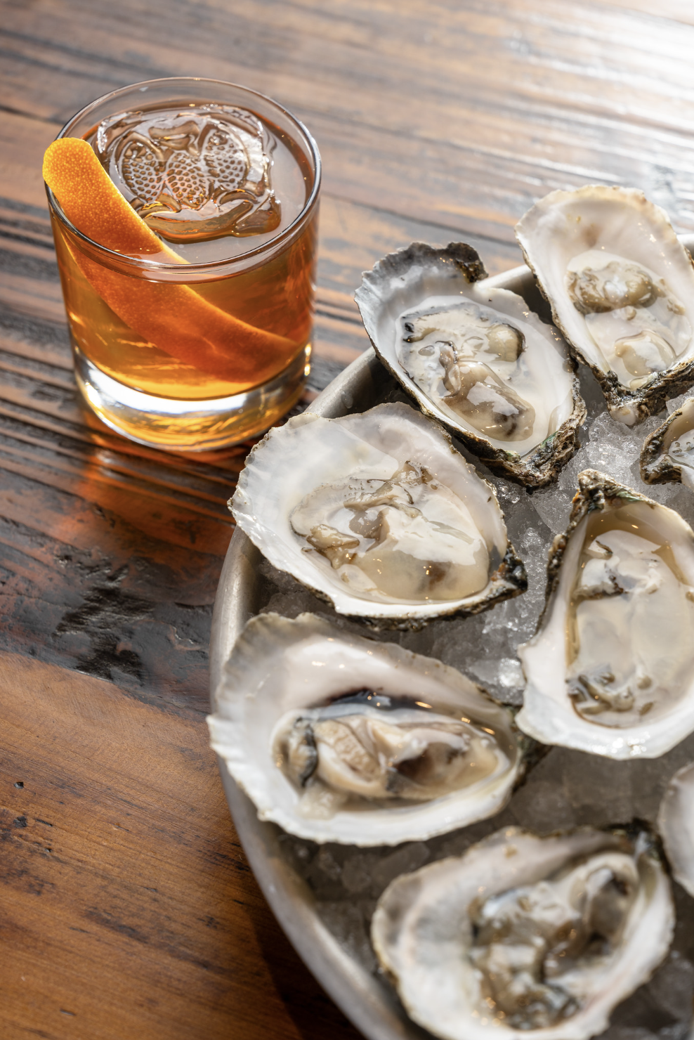 A glass of iced tea garnished with an orange slice next to a plate of fresh oysters on ice.