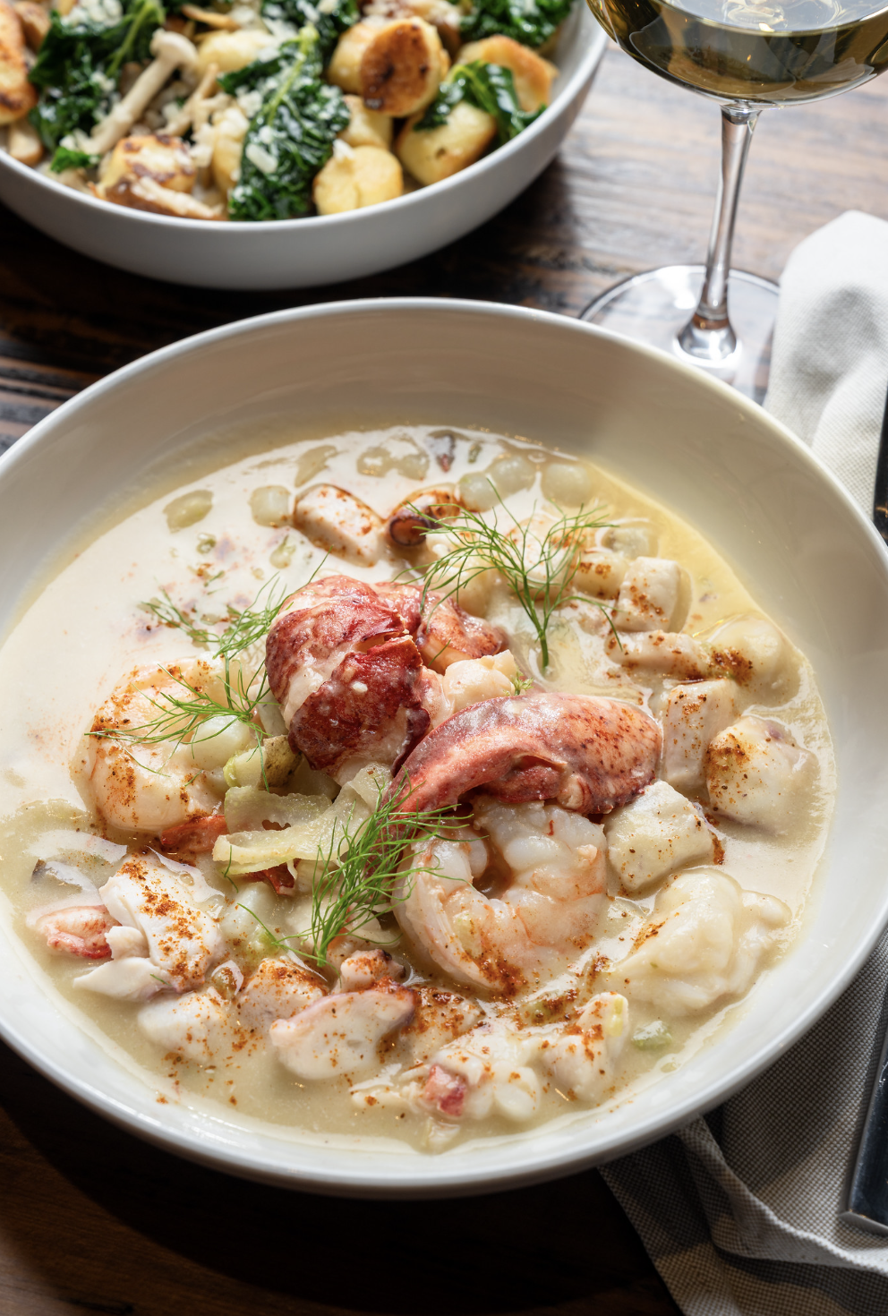 Seafood chowder with shrimp, lobster, and herbs in a white bowl, served with a glass of white wine and a side salad with greens and roasted potatoes.