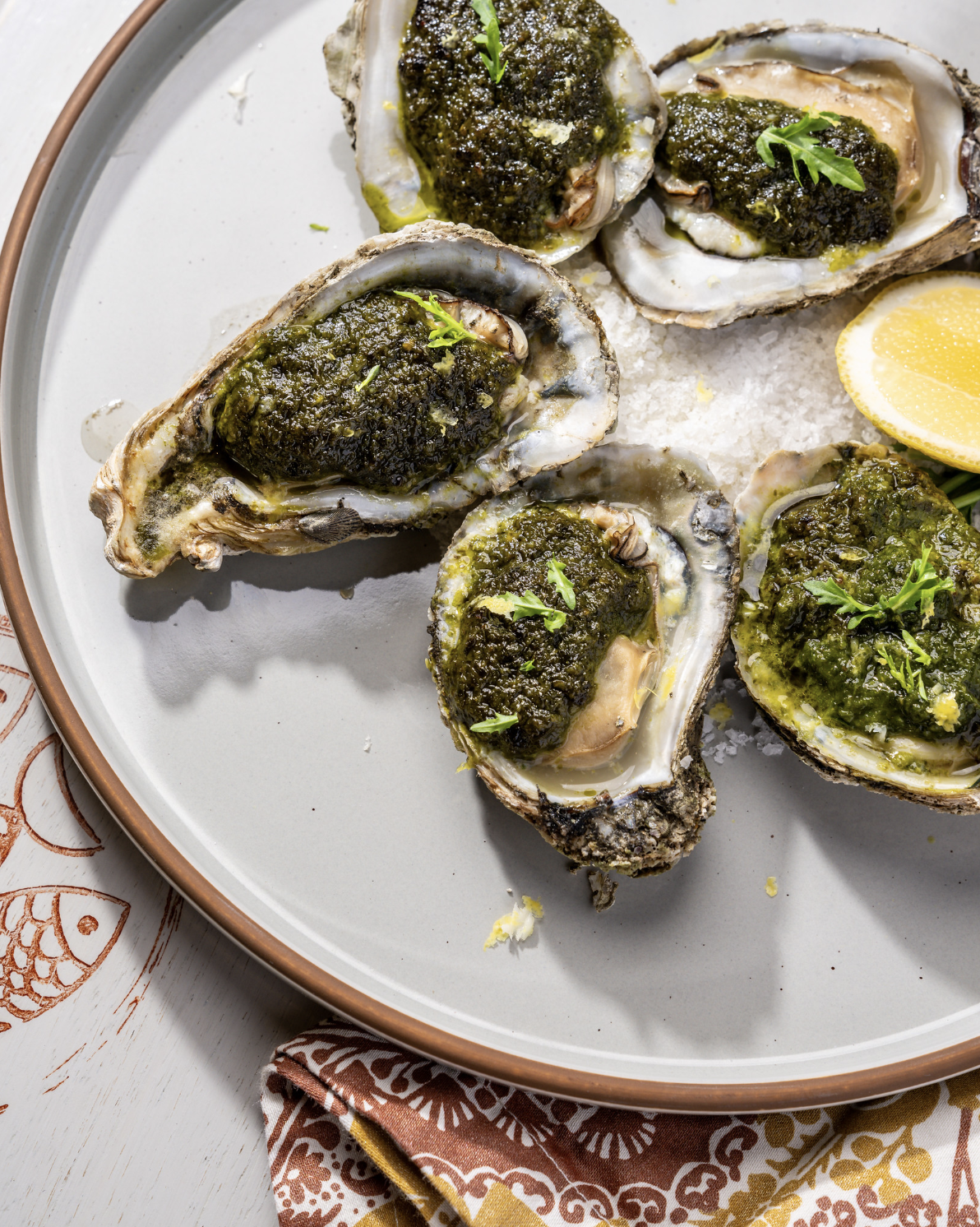 A plate of grilled oysters topped with green herbs and lemon wedges.