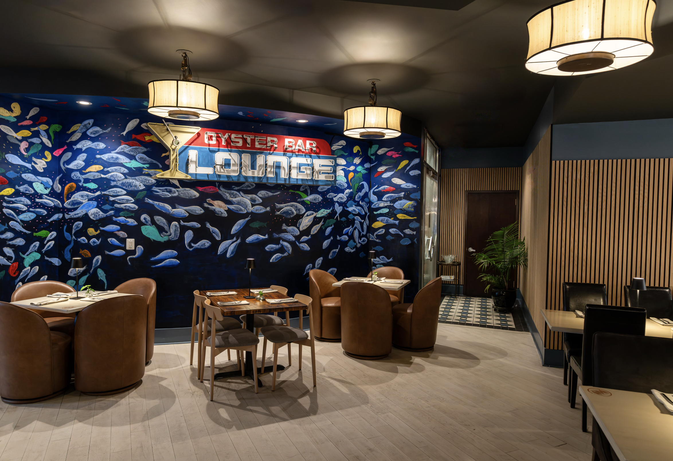 Interior of a seafood restaurant with a colorful fish mural, wooden tables and chairs, hanging ceiling lights, and a potted plant in the corner.