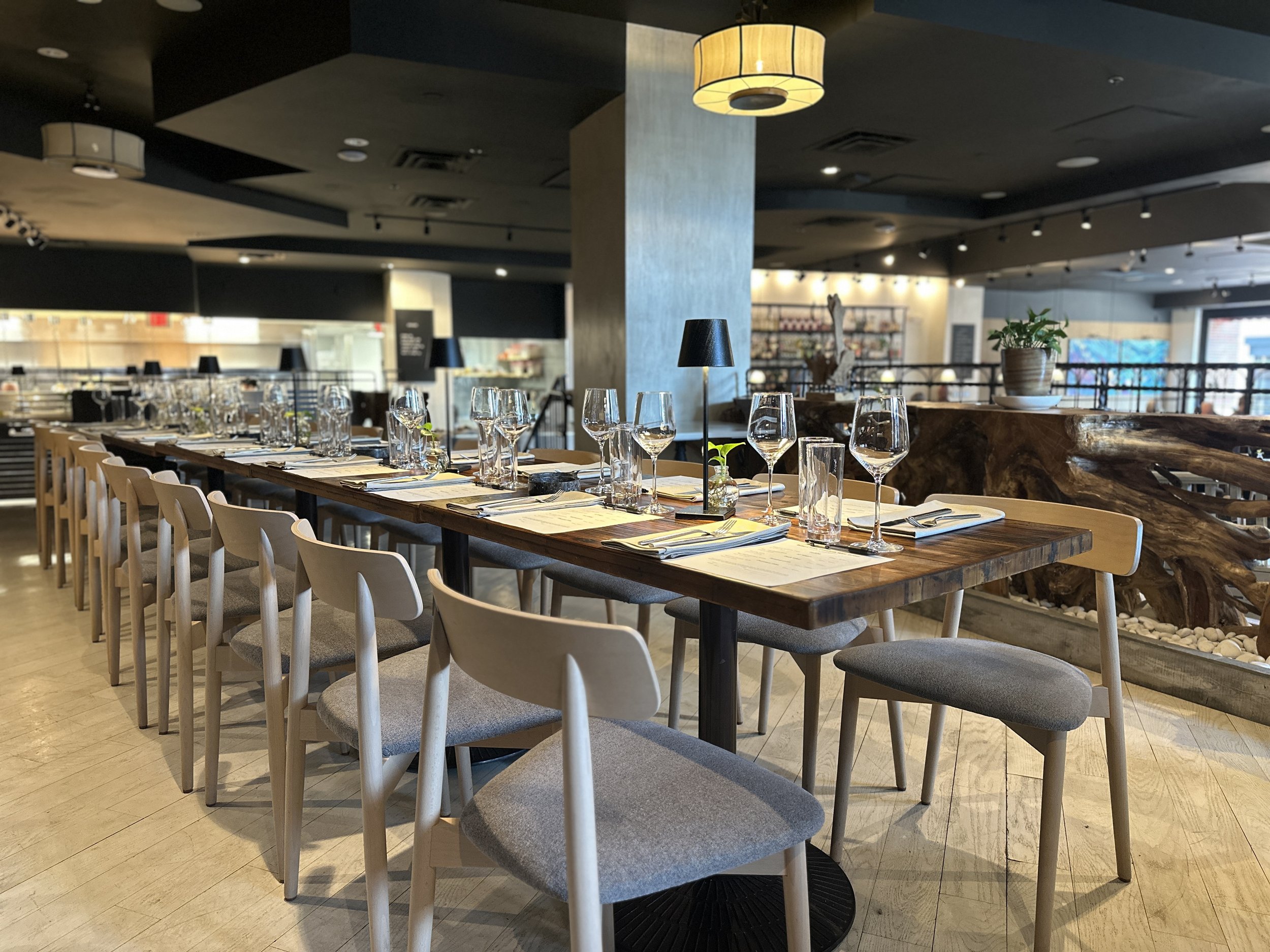 A restaurant interior with a long wooden table set with wine glasses, silverware, and menus. Light-colored chairs surround the table. In the background, there is a staircase, a potted plant on a stone countertop, and a store or display area.