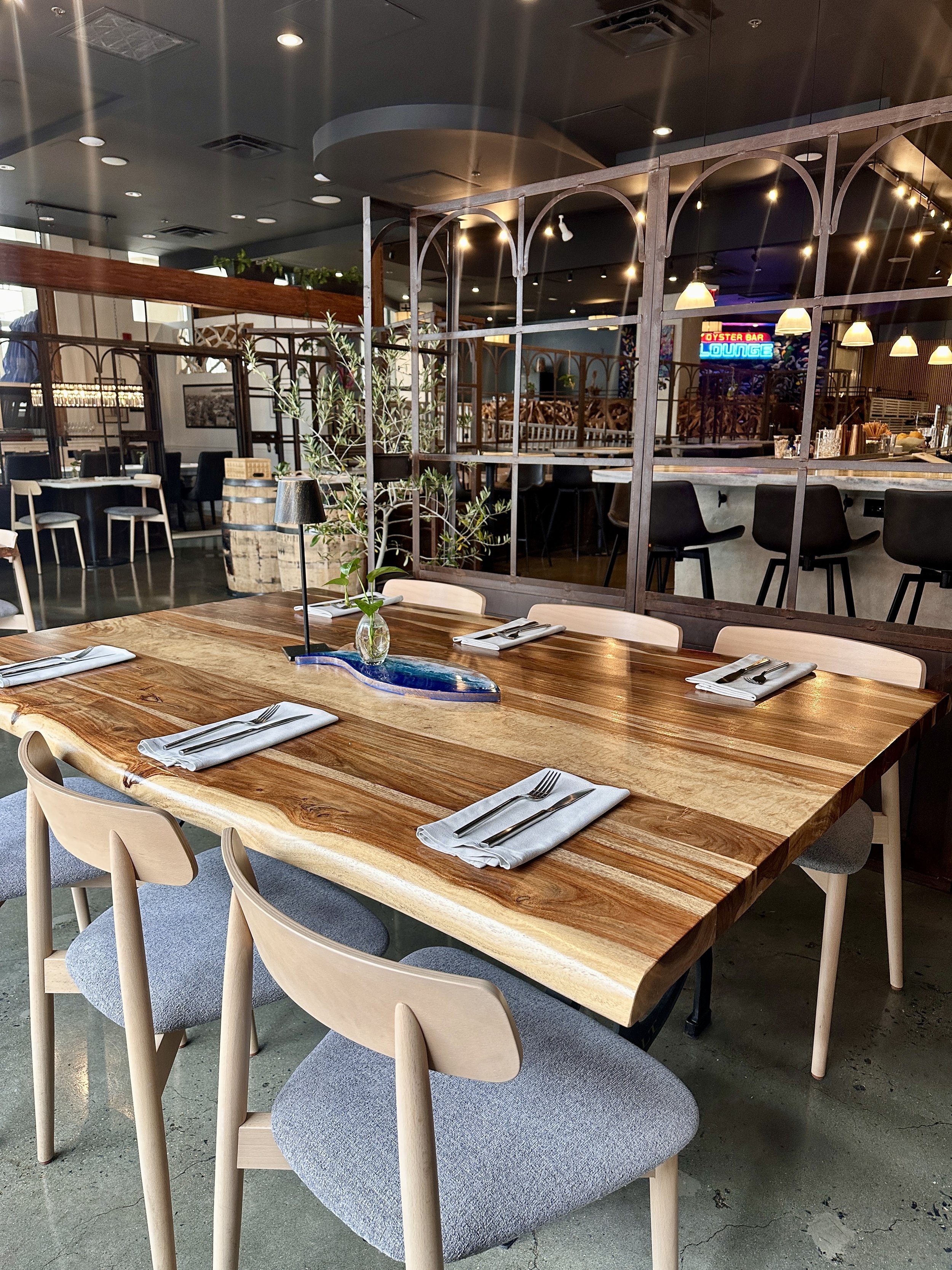 A wooden dining table set with white napkins, forks, and knives in a restaurant. The seating includes light-colored chairs with gray cushions. There is a small plant in a glass vase and a black table lamp on the table. In the background, there is a b