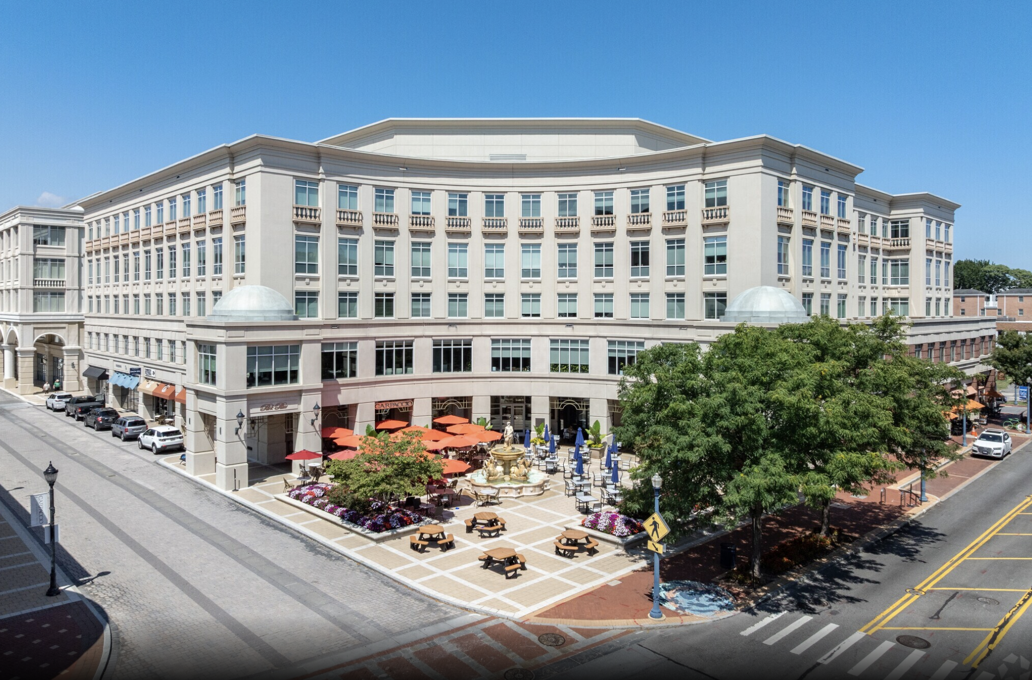 A large multi-story building with beige exterior and numerous windows, featuring a small outdoor seating area with orange umbrellas, a fountain, benches, trees, and colorful flower beds in front. The scene is set on a bright, sunny day with a clear blue sky.