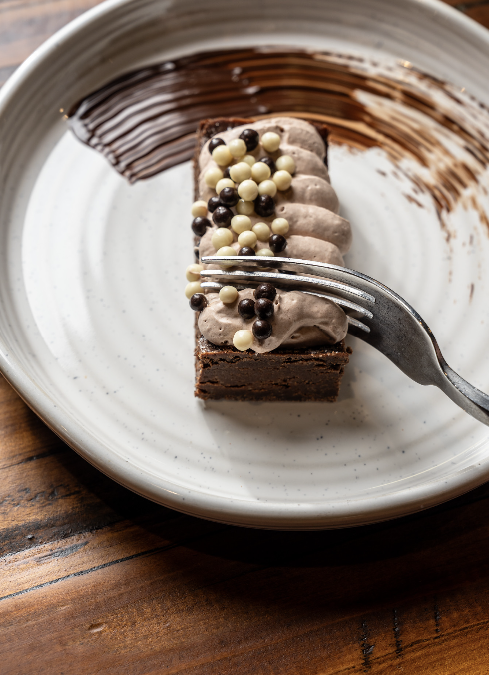 A slice of chocolate cake with chocolate frosting, topped with white and dark chocolate pearls on a white plate with chocolate sauce streaks, on a wooden table.