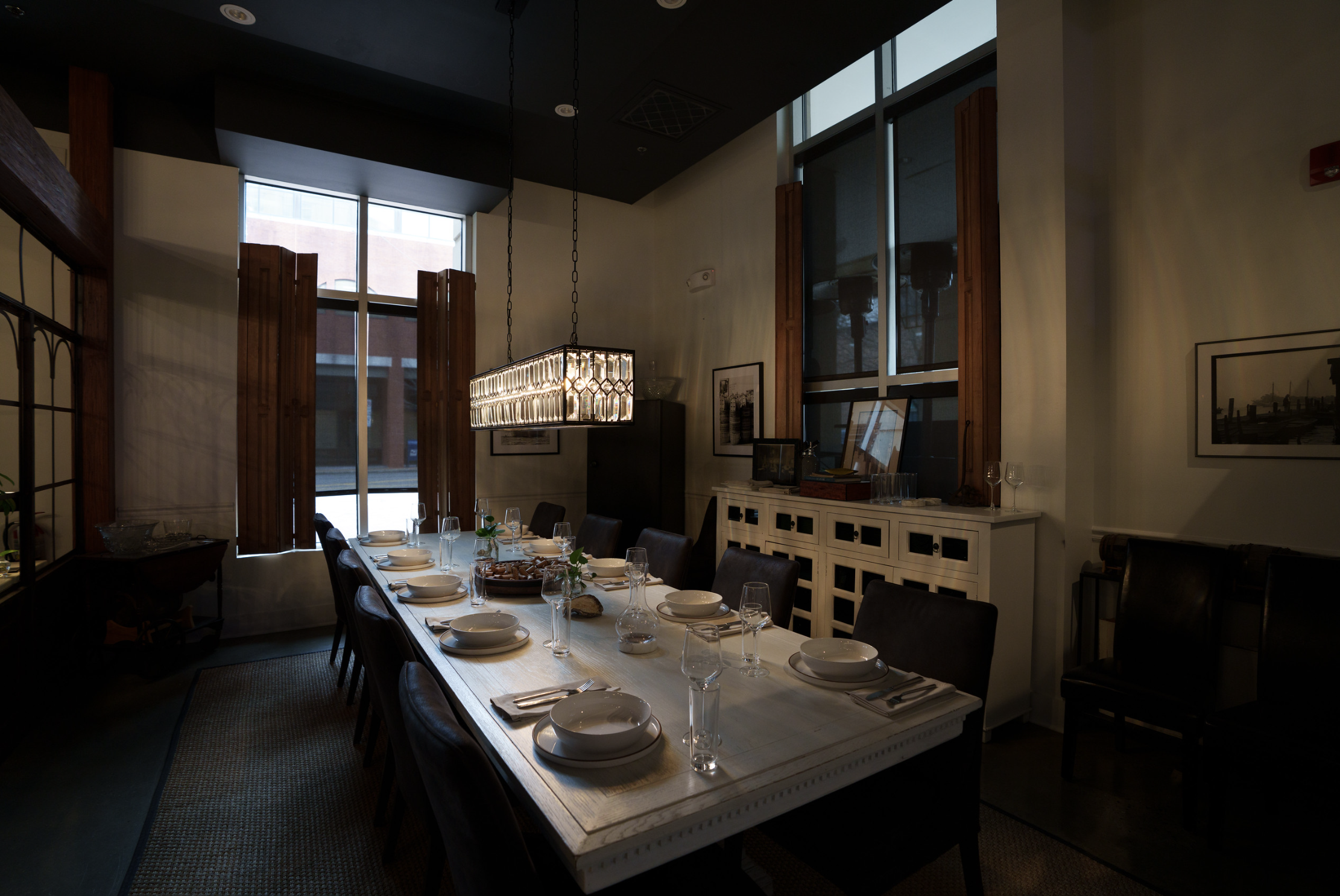 Dining room with a long table set with plates, glasses, and napkins, large windows with wooden frames, and a modern chandelier hanging above the table.