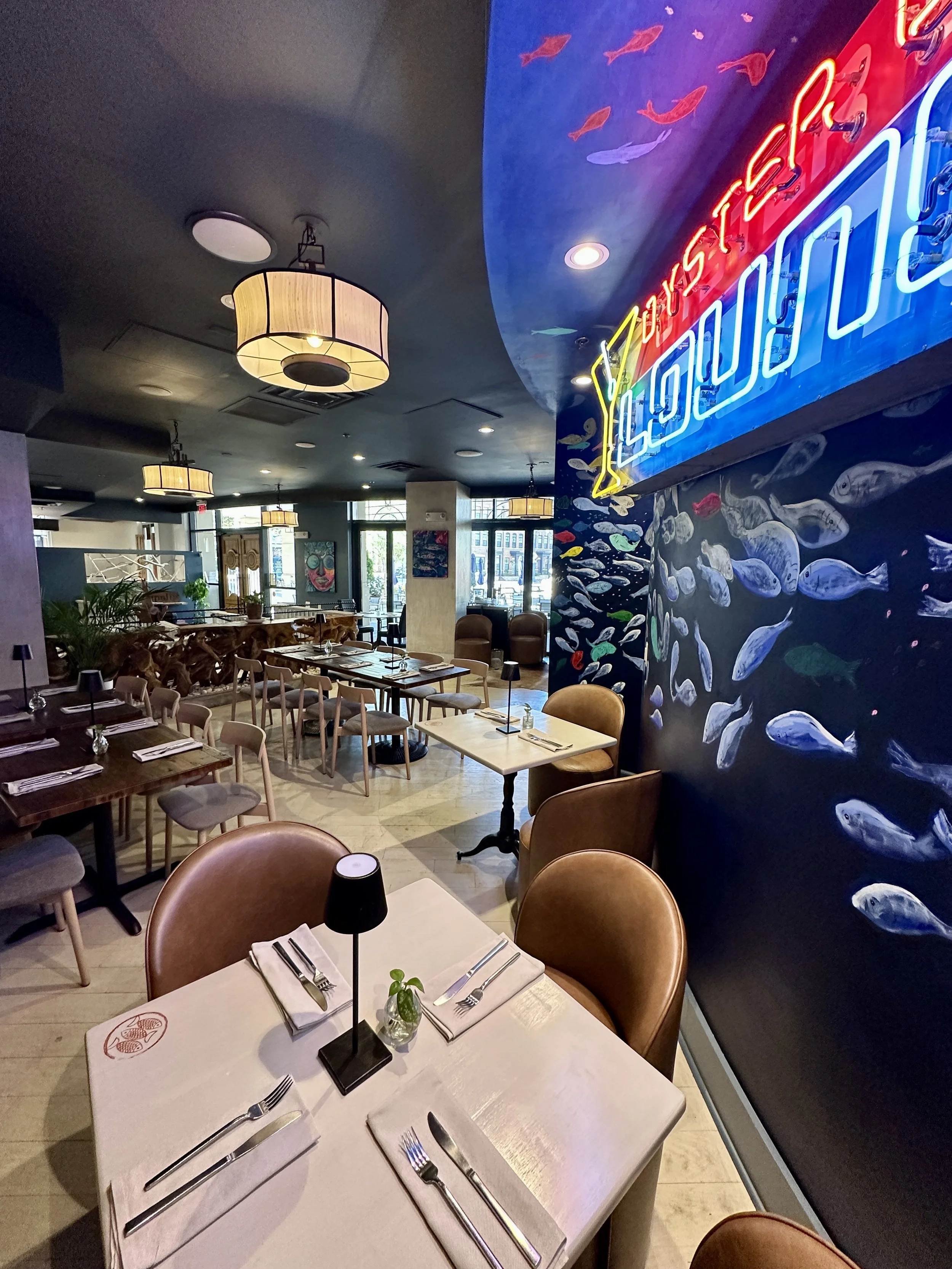 Interior of a restaurant with tables set for dining, featuring napkins, cutlery, small plant decorations, and modern ceiling lights. A colorful ocean-themed mural with fish and a neon sign with vibrant colors is visible on the wall.