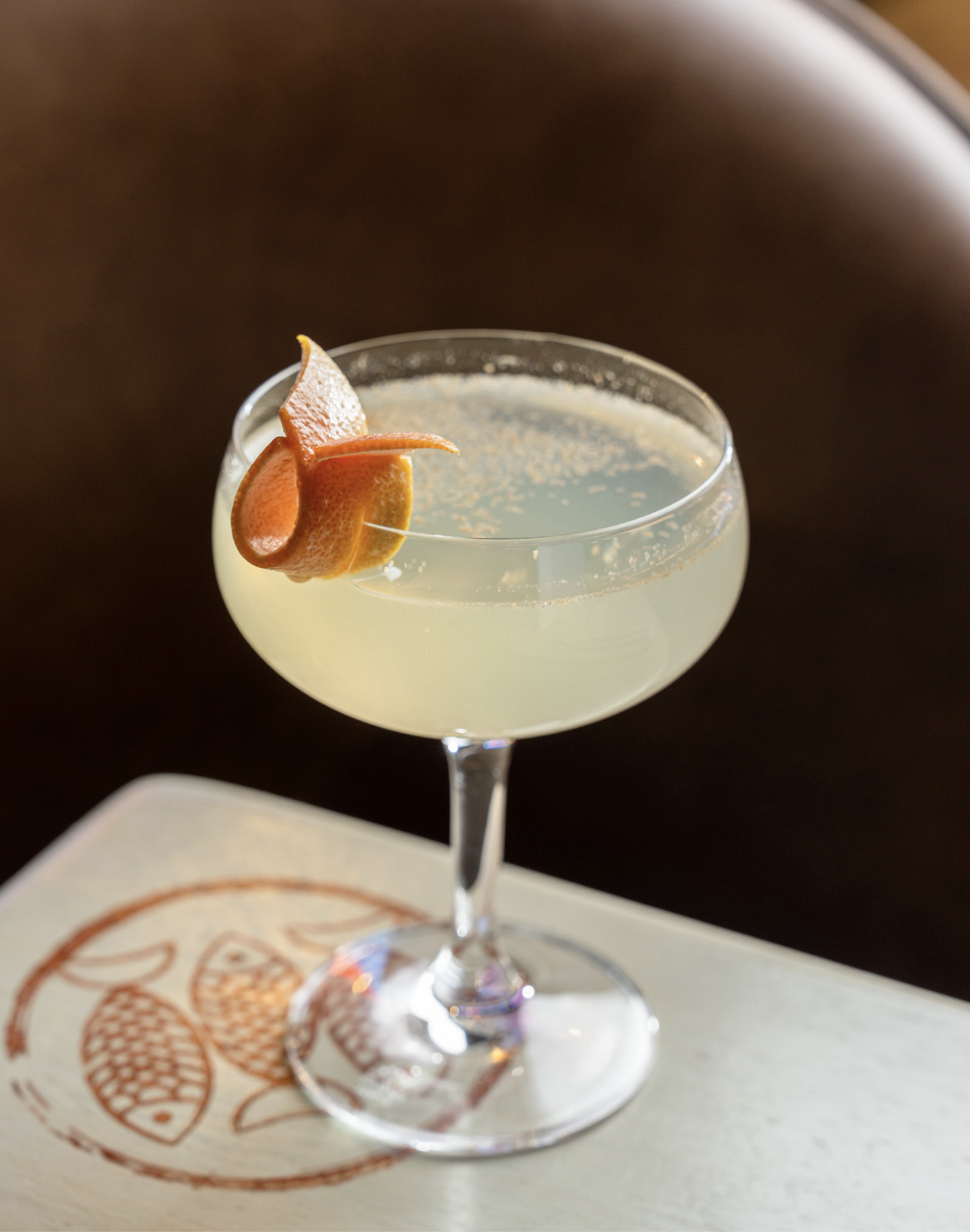 A cocktail in a coupe glass garnished with orange peel on a white napkin with a stylized fish design.