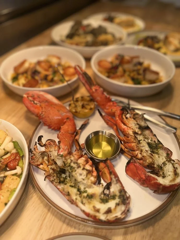 Lobster tail halves with melted cheese and herbs, served with dipping oil, on a white plate with side dishes of pasta and seafood in the background.
