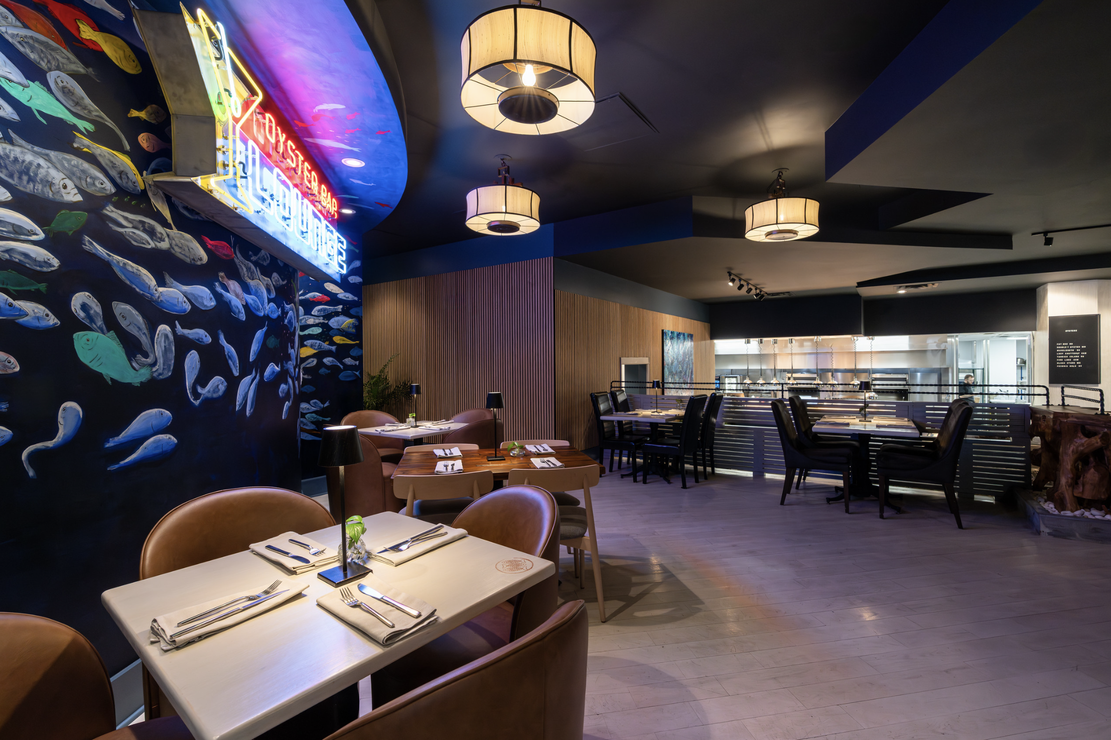 Upscale restaurant interior with pastel-colored tables, leather chairs, wall art featuring fish, and neon signs, illuminated by large hanging pendant lights.