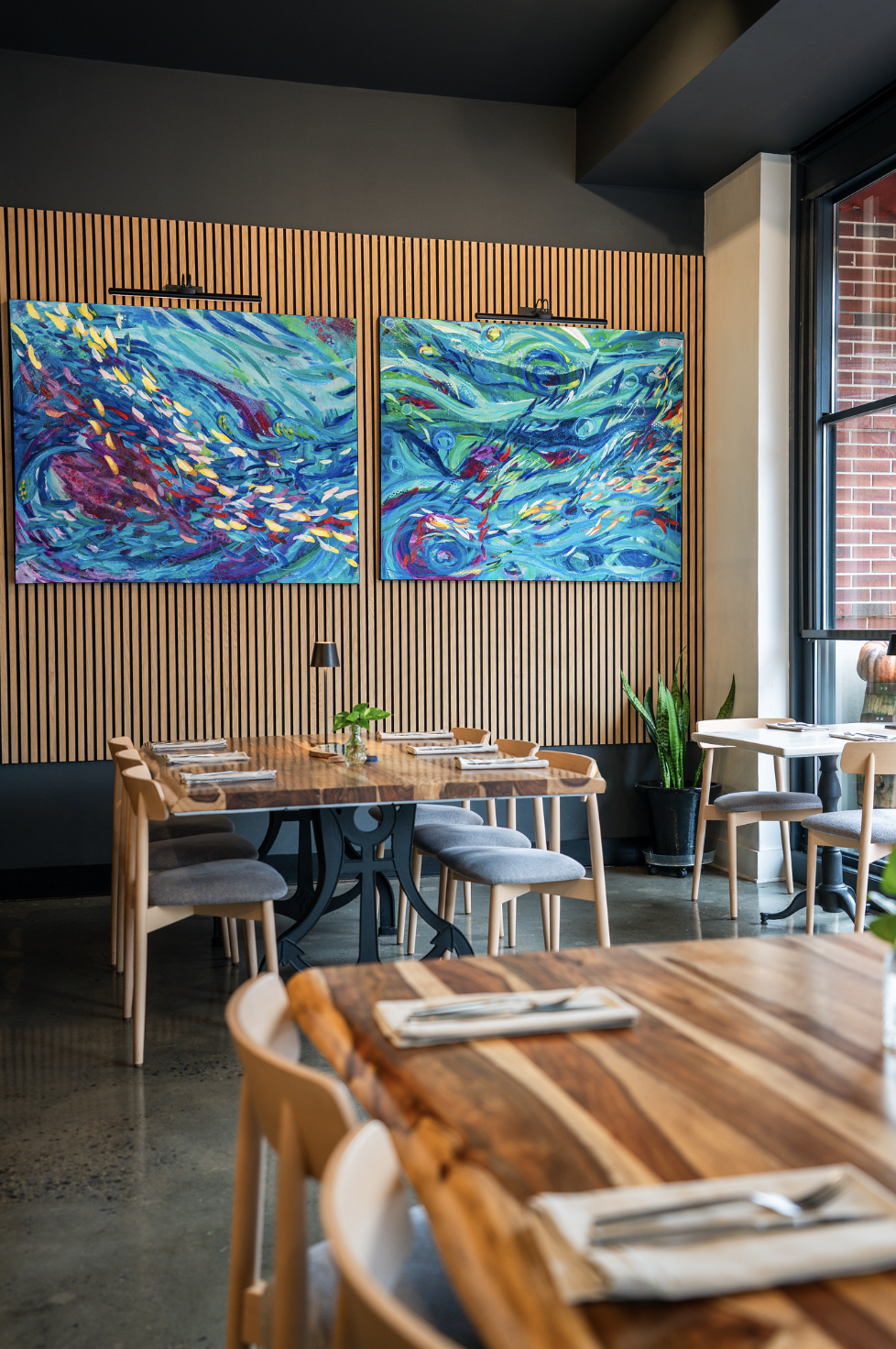 Interior of a modern restaurant with wooden tables, gray chairs, green plants, and colorful abstract paintings on a wooden-paneled wall near large windows.