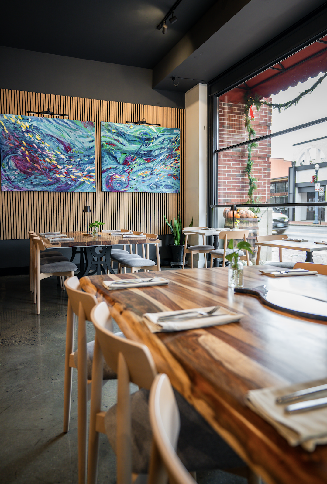 Interior of a modern restaurant with wooden tables, light-colored chairs, and colorful abstract paintings on the wall. Large window with holiday decorations and potted plants.