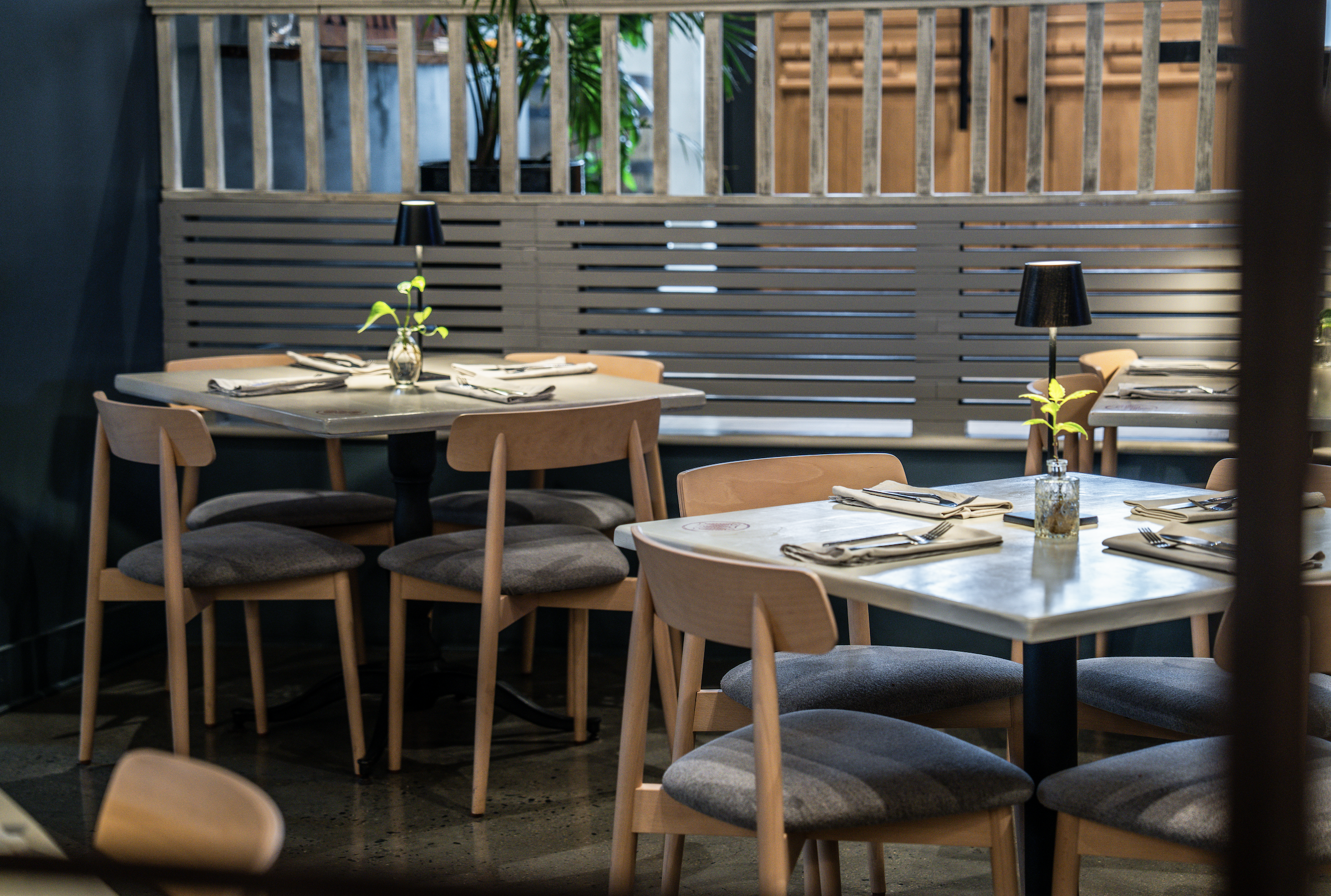 Interior of a modern restaurant or cafe with wooden chairs, tables set with napkins and utensils, small vases with green plants, and black lamps, with a wooden partition and some greenery outside.