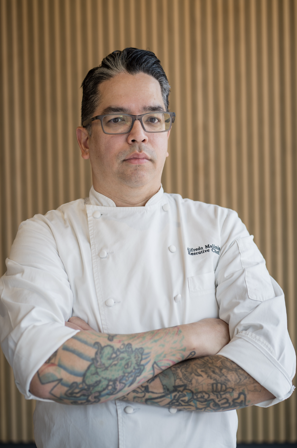 A professional chef with glasses and tattoos on his arms, standing with crossed arms in front of a beige curtain, wearing a white chef's coat.
