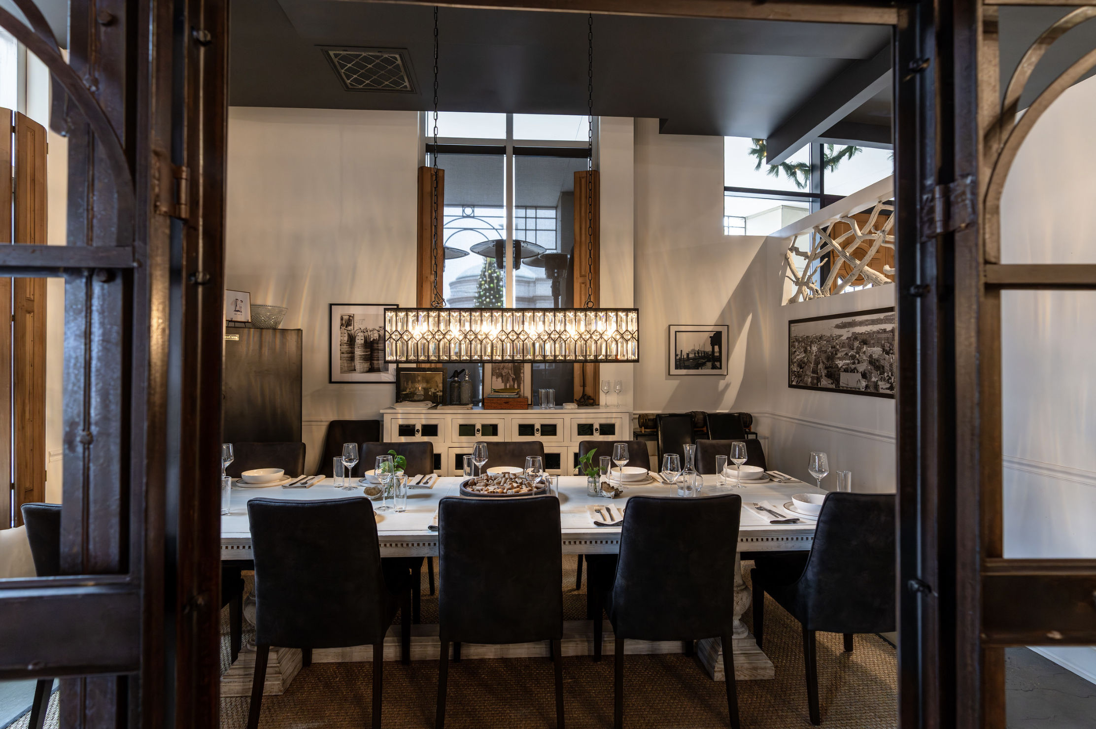 A dining room with a long table set with glasses, plates, and cutlery, arranged for a meal, and a modern chandelier hanging overhead.