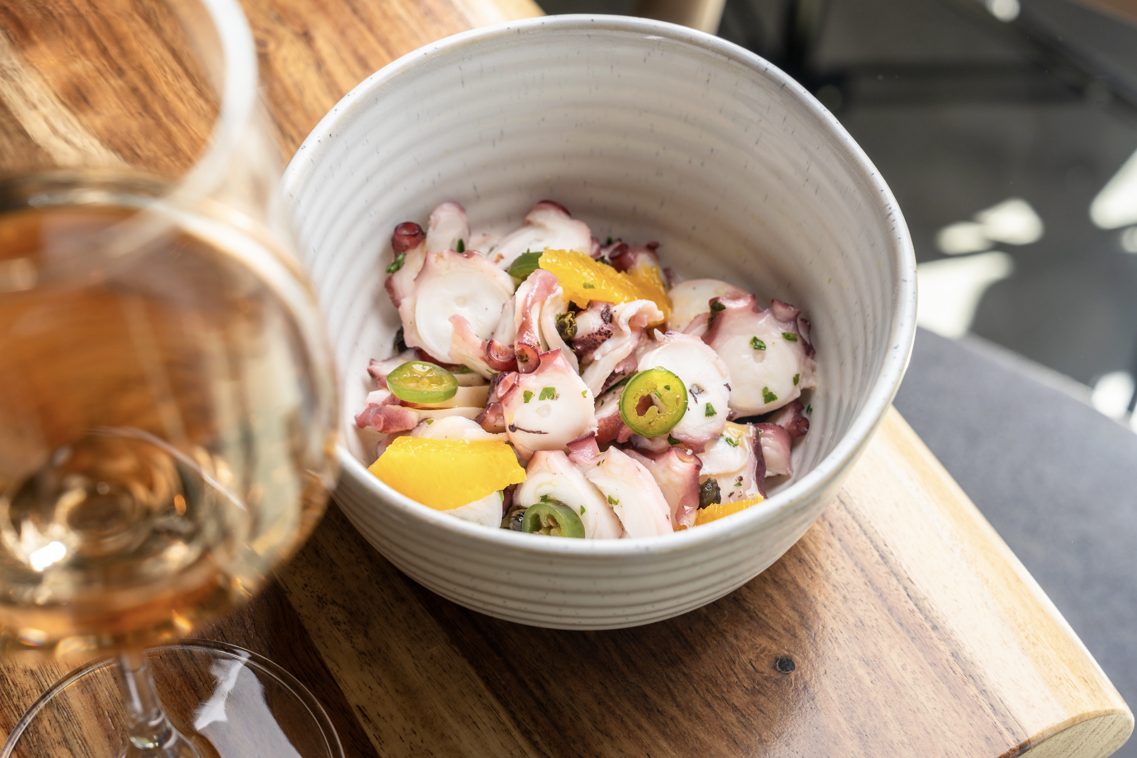 A white bowl containing octopus salad garnished with green chili slices, yellow bell pepper slices, and chopped herbs, placed on a wooden surface beside a glass of rosé wine.