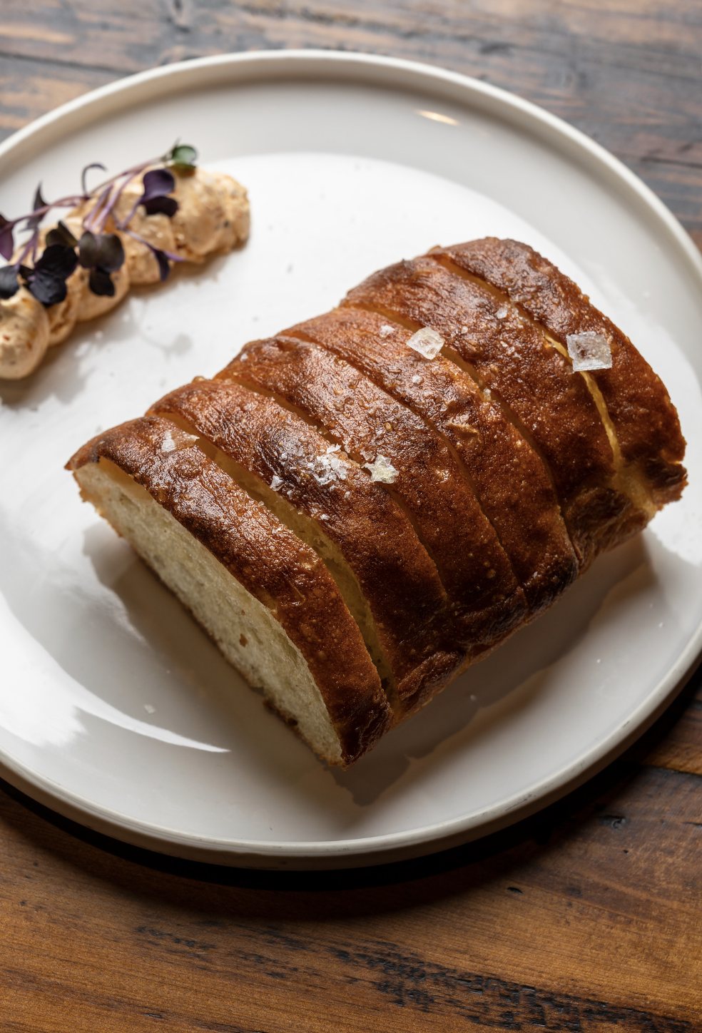 Sliced loaf of bread on a white plate with salt crystals on top, accompanied by a side of creamy spread topped with microgreens, on a wooden table.