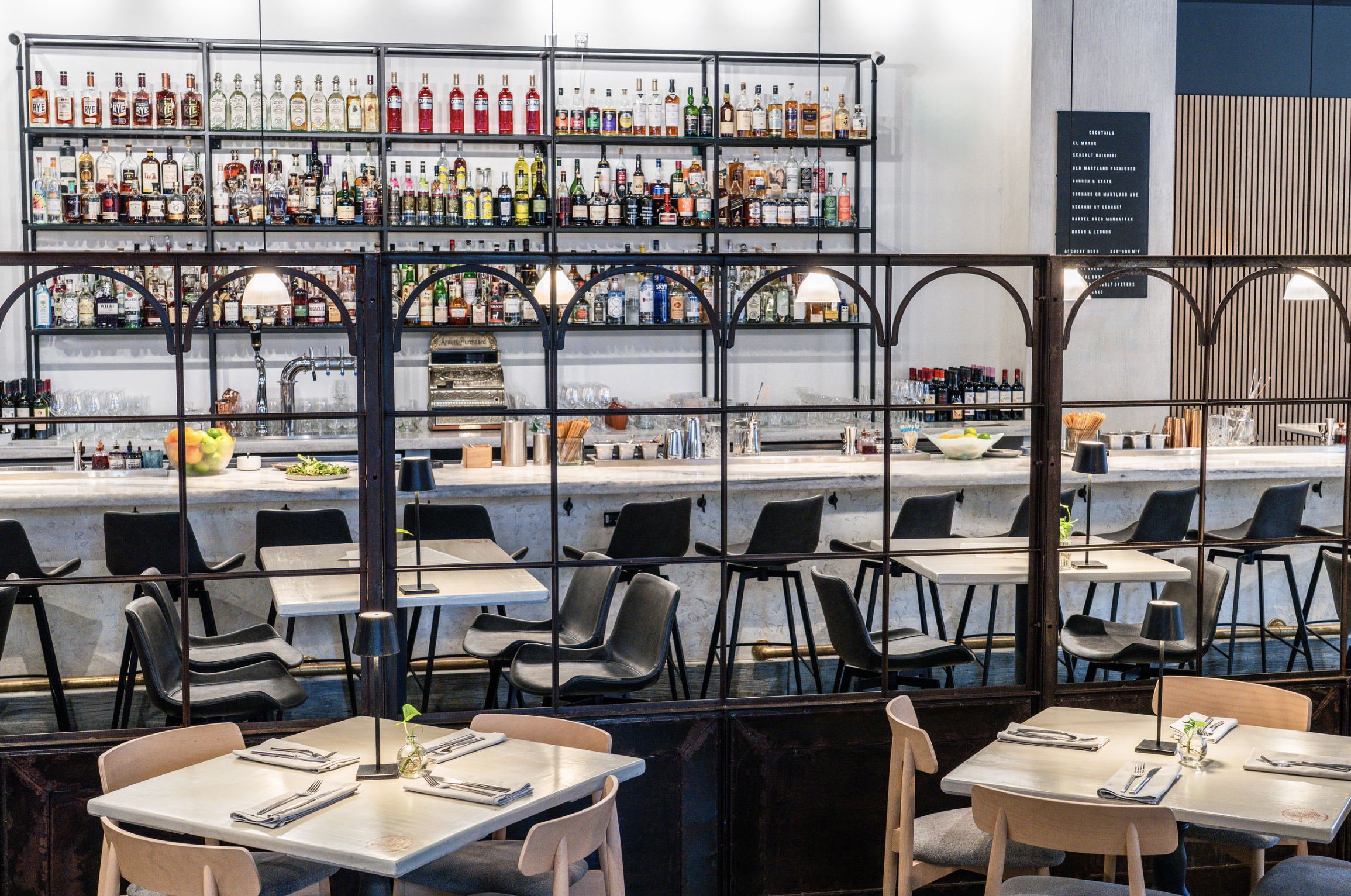 Interior of a bar and restaurant with a well-stocked shelves of liquor bottles behind the bar and tables set with napkins, glasses, and utensils in the seating area.