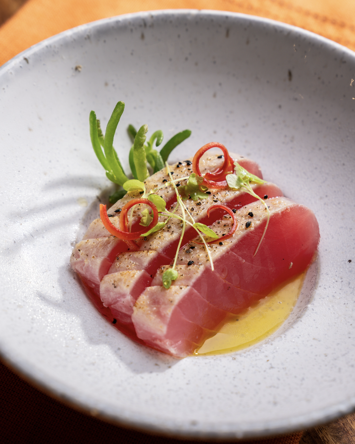 Seared tuna sashimi with microgreens, red chili pepper slices, and black sesame seeds on a white plate.