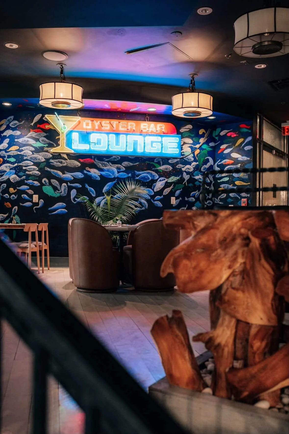 Interior of a restaurant featuring a neon sign that reads 'Oyster Bar Lounge,' decorated with a fish-themed mural, hanging ceiling lights, and a wooden sculpture in the foreground.