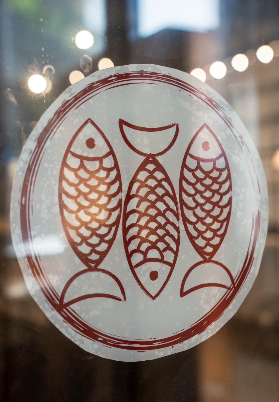 Sticker of three fish arranged vertically, with a moon shape above the middle fish, on a glass window with city lights reflected behind.