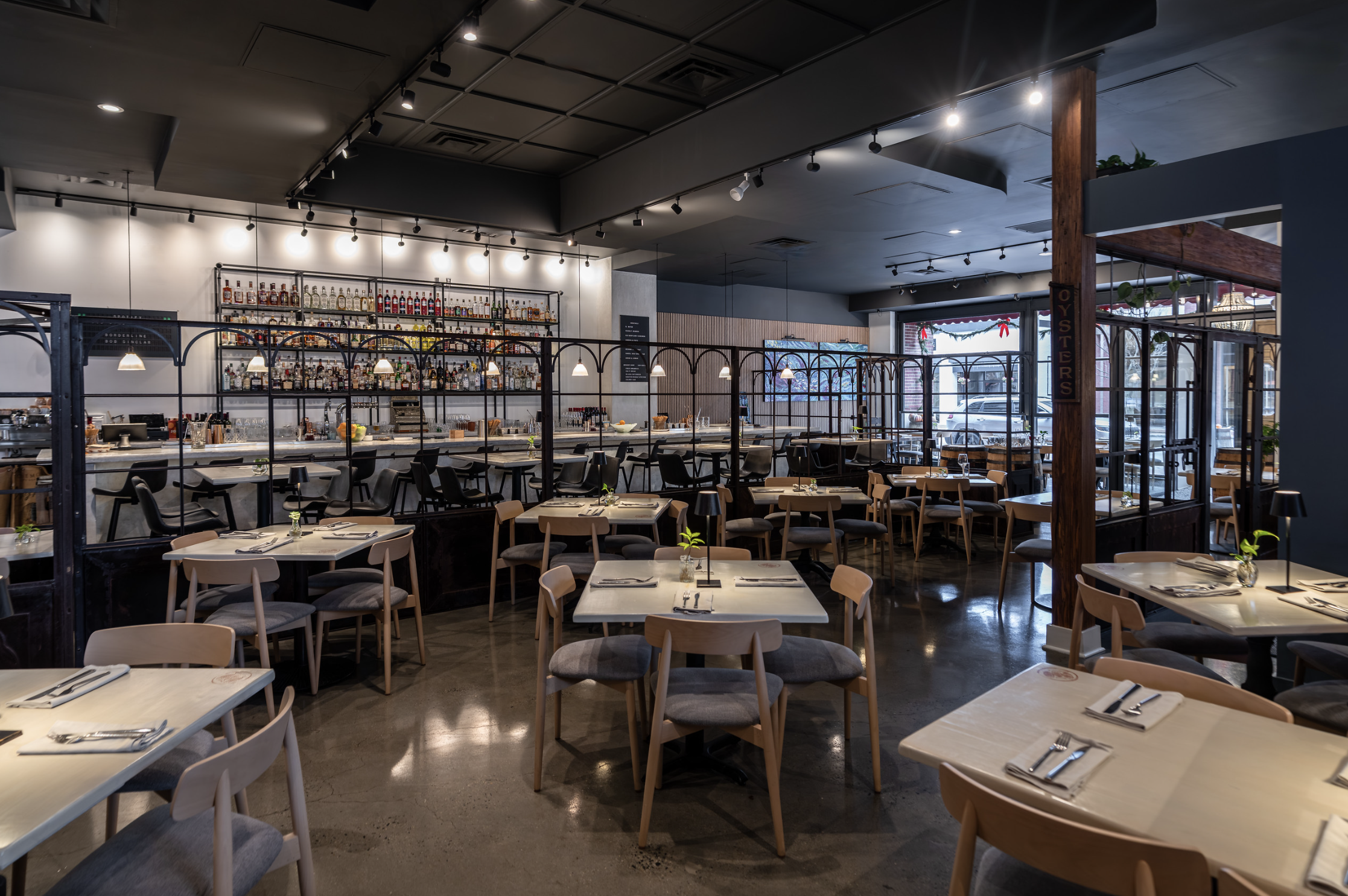 Empty restaurant with neatly set tables, bar area with liquor bottles behind, large windows, modern decor with black and wood accents.
