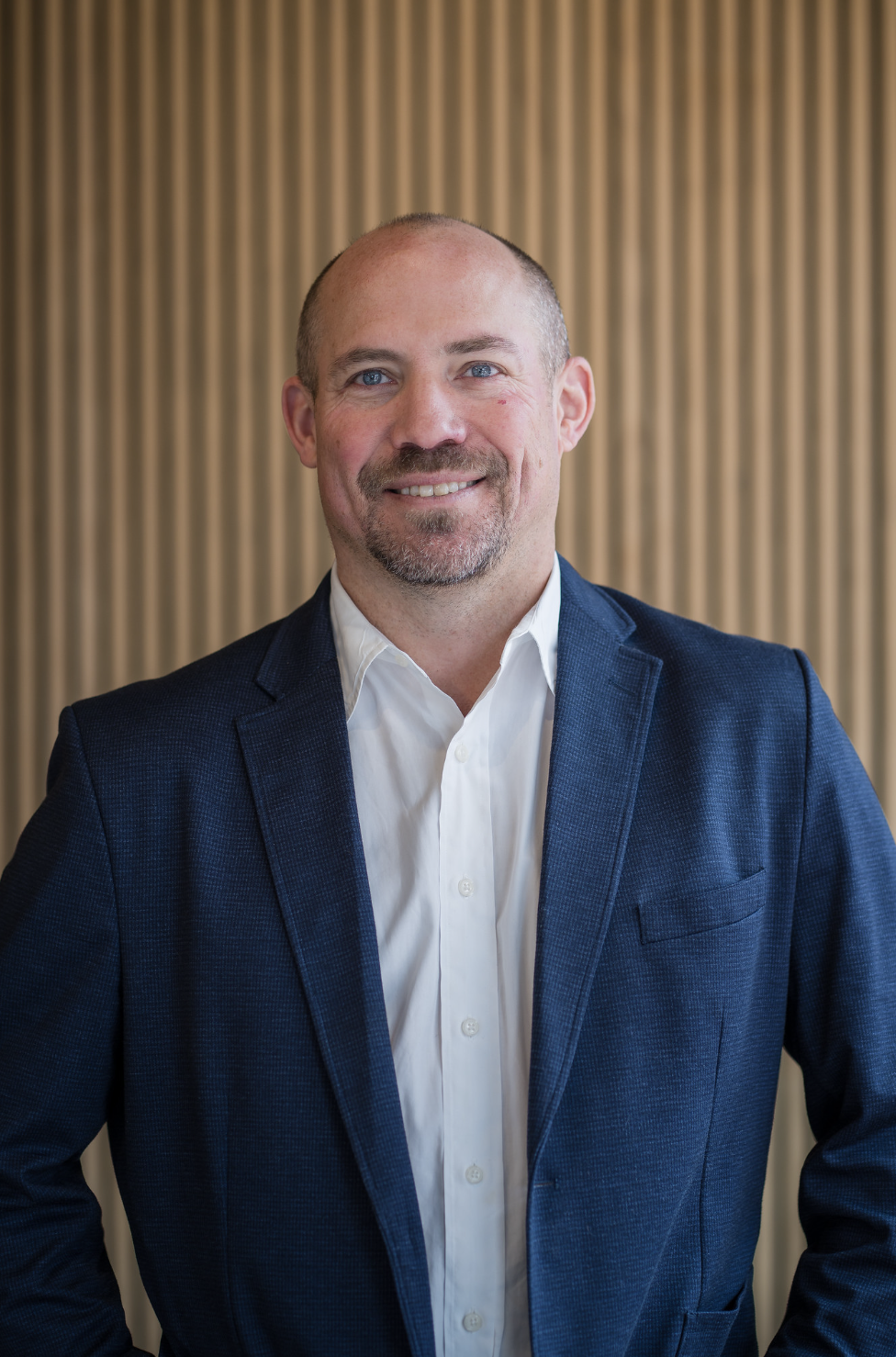 Portrait of a man with a friendly smile, wearing a navy blazer and white shirt, standing against a textured beige background.