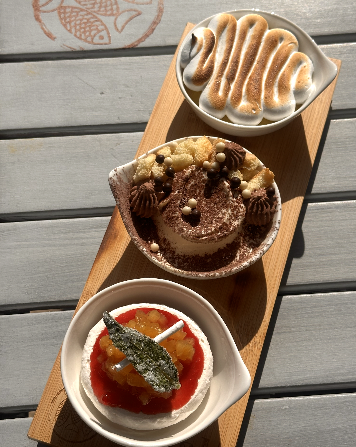 Three bowls of desserts on a wooden tray: a bowl of toasted marshmallows in cream, a bowl of tiramisu topped with chocolate and white balls, and a bowl of fruit-topped panna cotta with a candied green leaf.