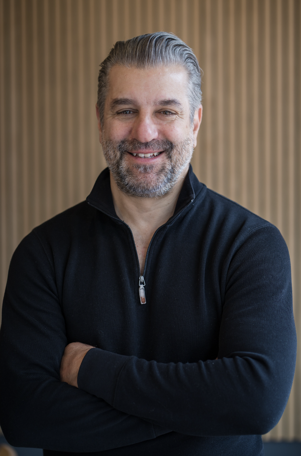 A middle-aged man with gray hair and a beard, smiling, wearing a black zip-up sweater with arms crossed, standing in front of a wooden slatted wall.