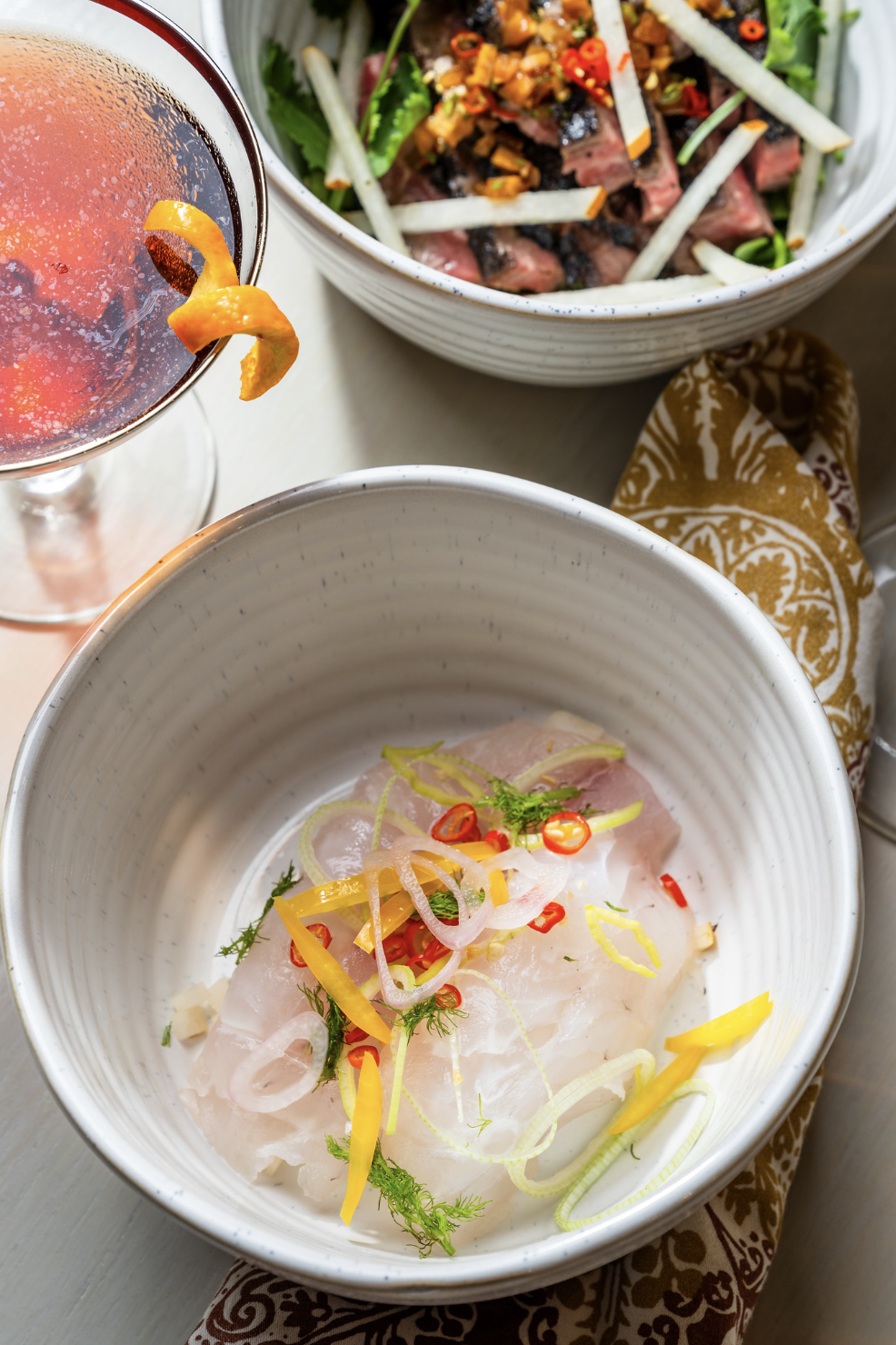 A bowl of raw fish garnished with sliced red chili, lemon zest, fennel, and green herbs, a salad with beef slices, green vegetables, and spicy toppings, and a glass of cocktail with lemon peel.