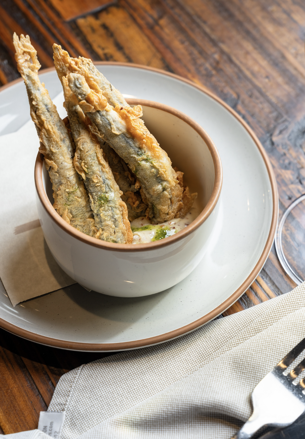 Fried fish fillets served in a white bowl on a beige plate, garnished with sauce and herbs, set on a wooden table with a fork and napkin nearby.