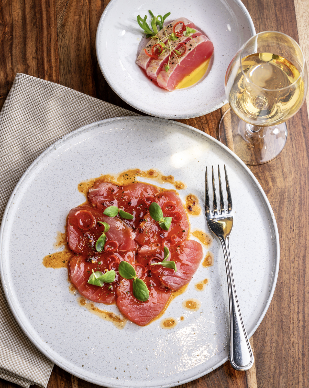 A plate of thinly sliced tuna carpaccio garnished with microgreens and red chili slices, served with a drizzle of sauce. A fork rests on the edge of the plate. In the background, a small dish contains more tuna slices with similar garnishes. A glass of white wine is also visible on a wooden table, with a beige napkin beside the plate.