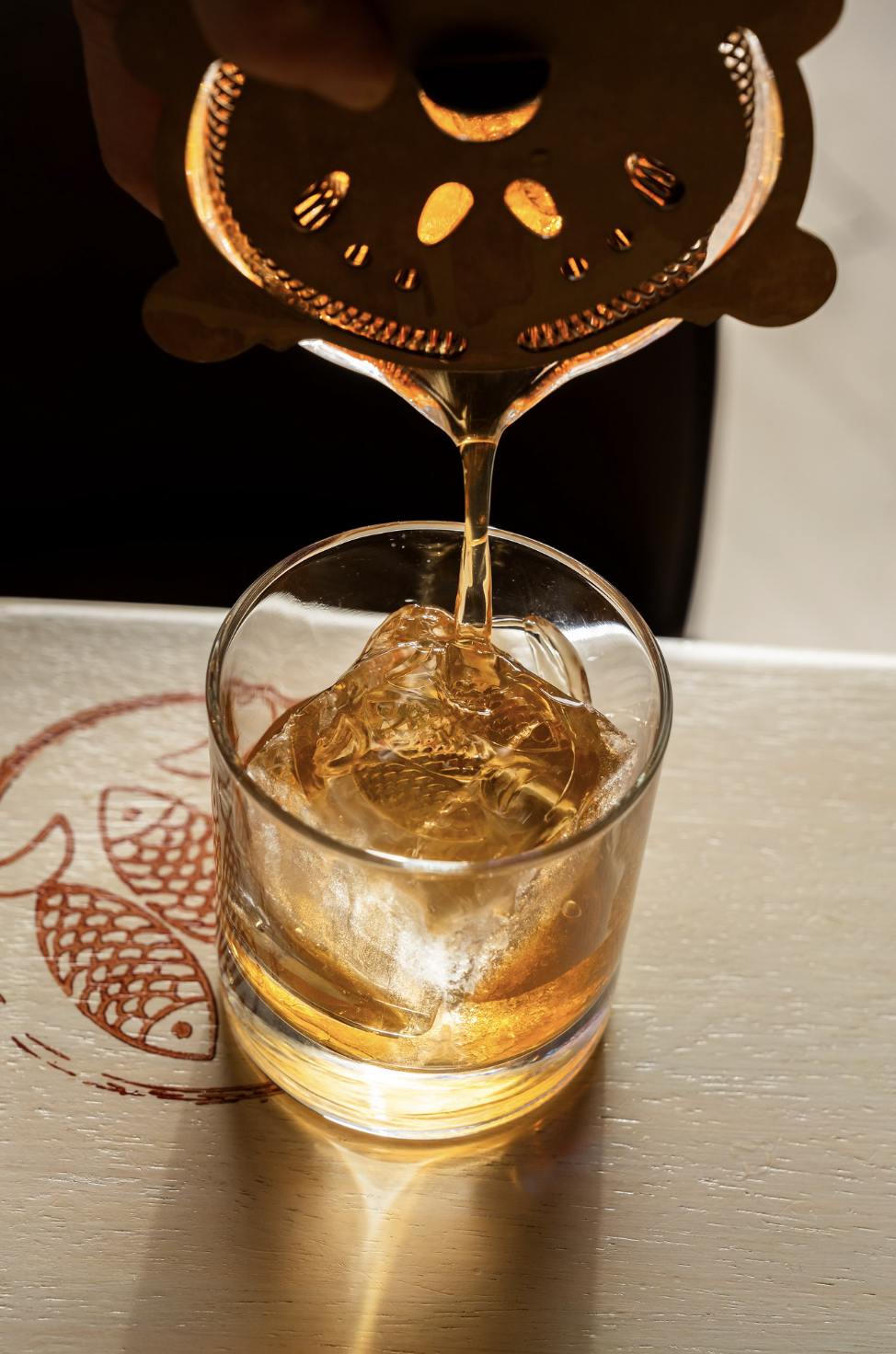 A hand places a metal strainer over a glass with ice and amber-colored liquid, capturing the pouring liquid.