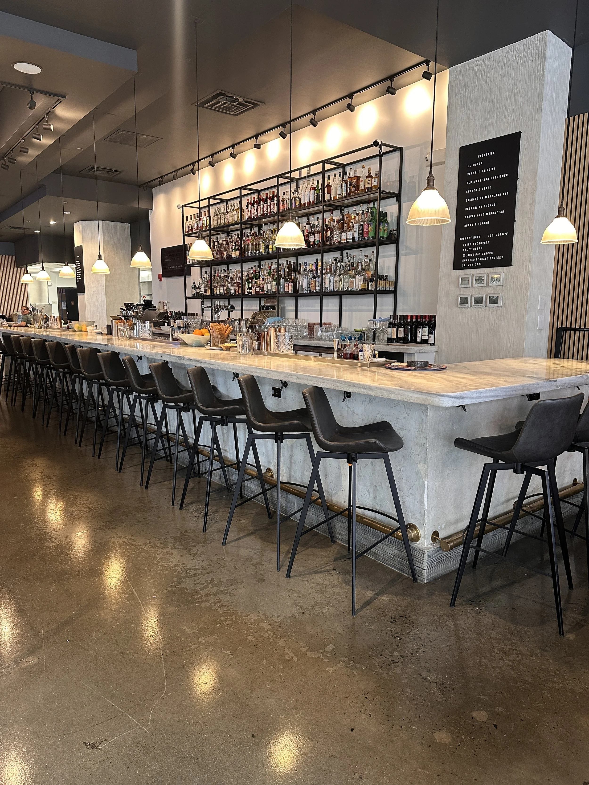 Inside a modern bar with a long marble countertop, black bar stools, and a large black metal shelf filled with various bottles of alcohol. There are hanging pendant lights over the bar and a black menu board on the wall listing cocktails.