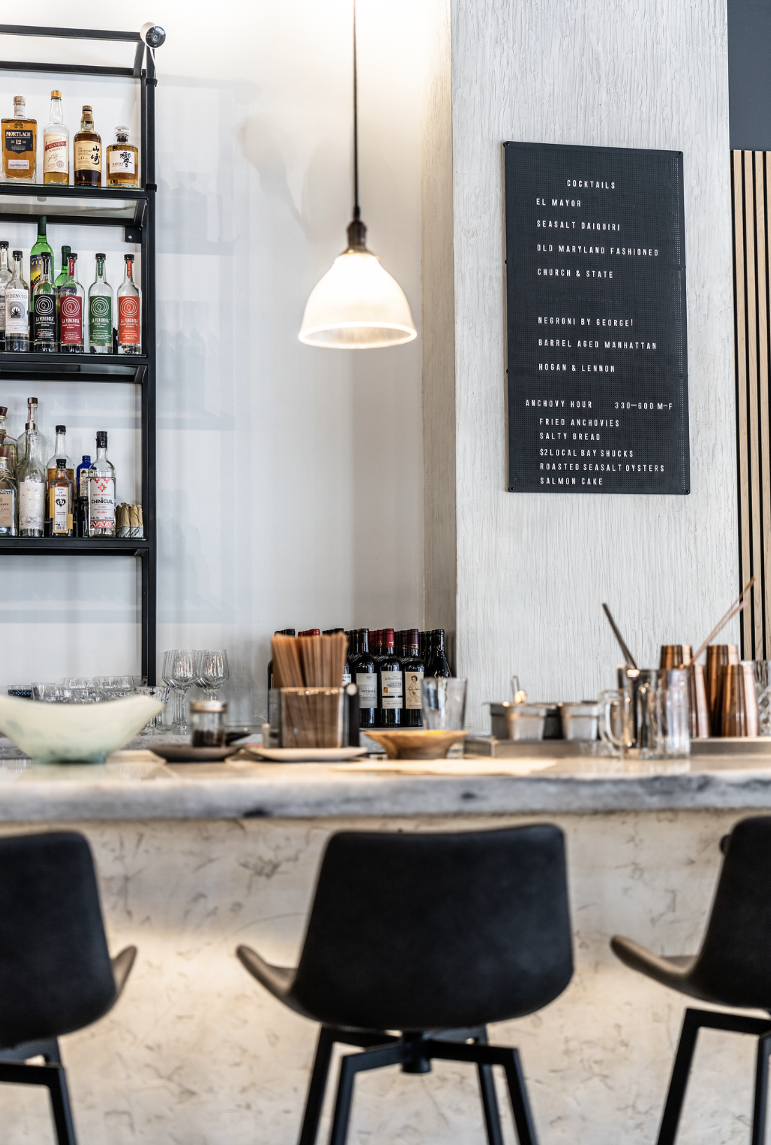 A bar with a black metal shelf holding various bottles of liquor, a chalkboard menu listing cocktail options, and a marble countertop with glasses, wine bottles, and bar tools. There are black bar stools in front of the counter.
