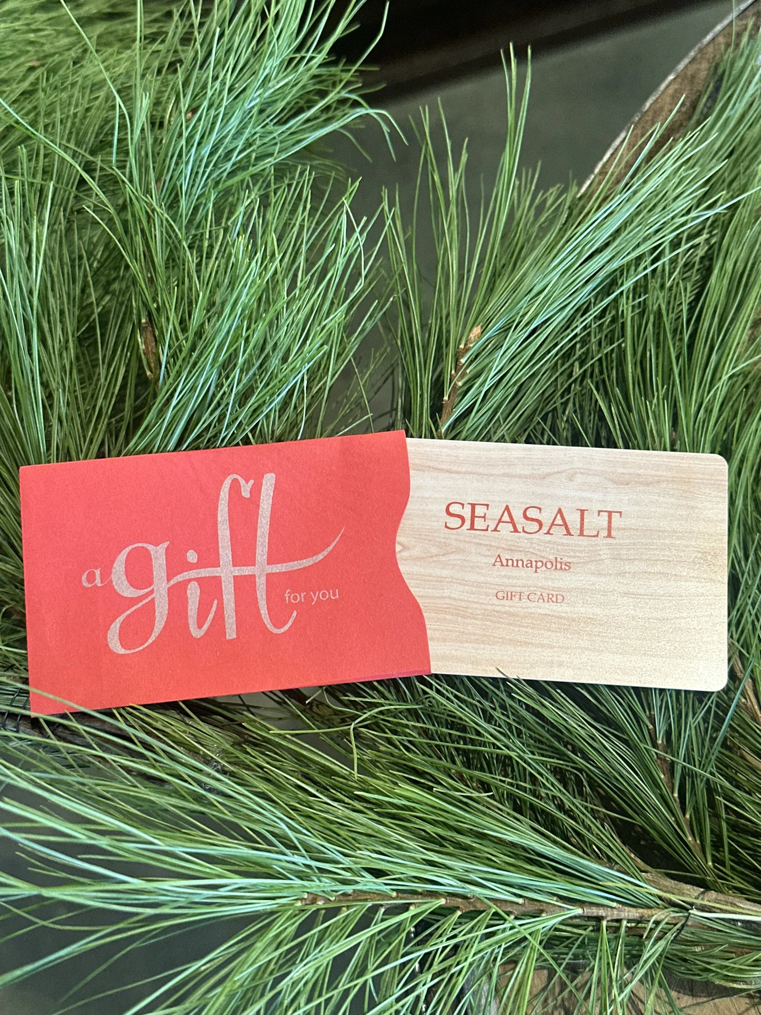 Green pine branches with a red and beige gift card that reads 'a gift for you' and 'Seasalt Annapolis'.