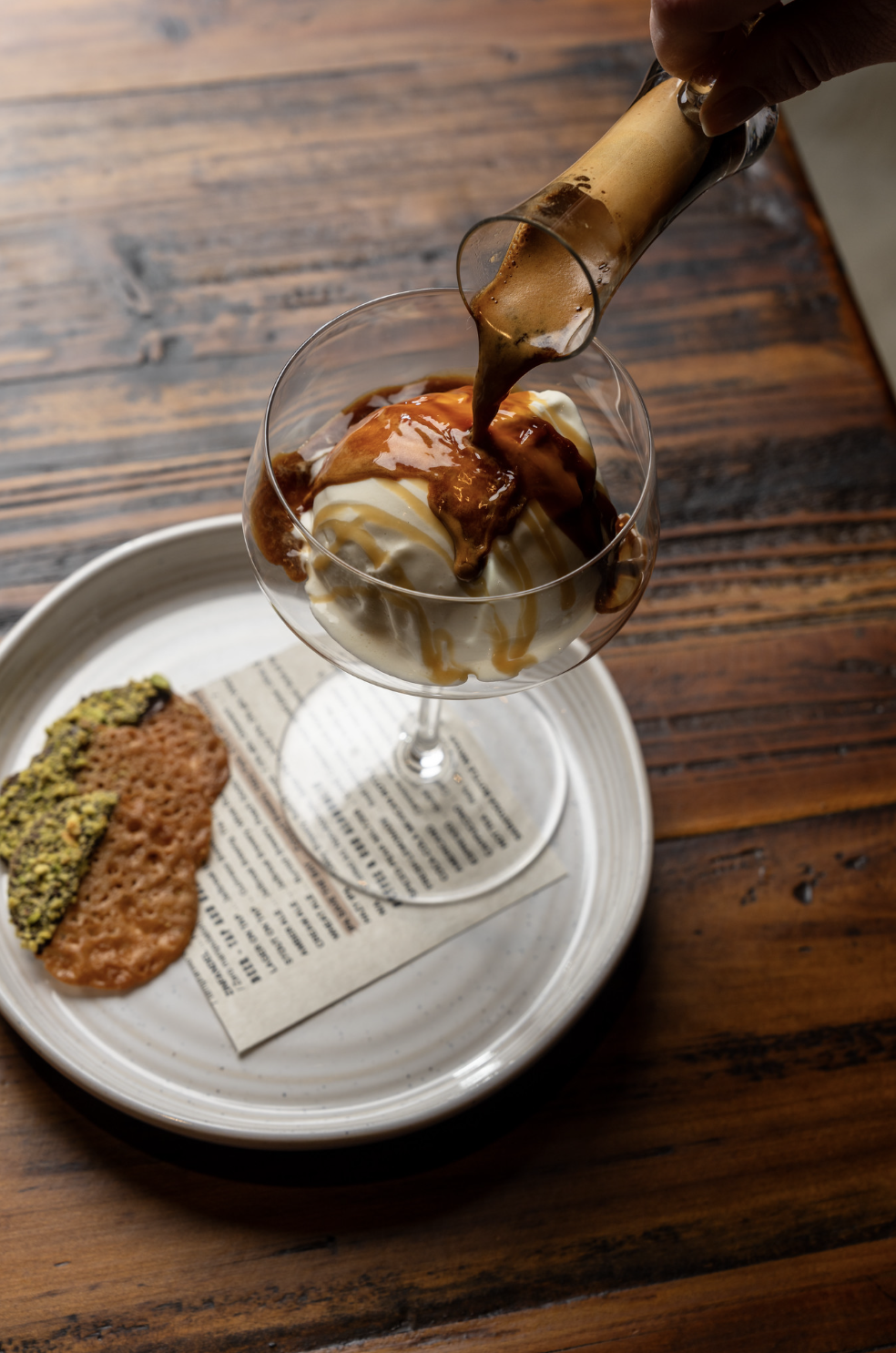 A dessert of vanilla ice cream in a glass with caramel sauce on top, being poured from a bottle, served on a white plate with a pistachio-topped pastry on a napkin.