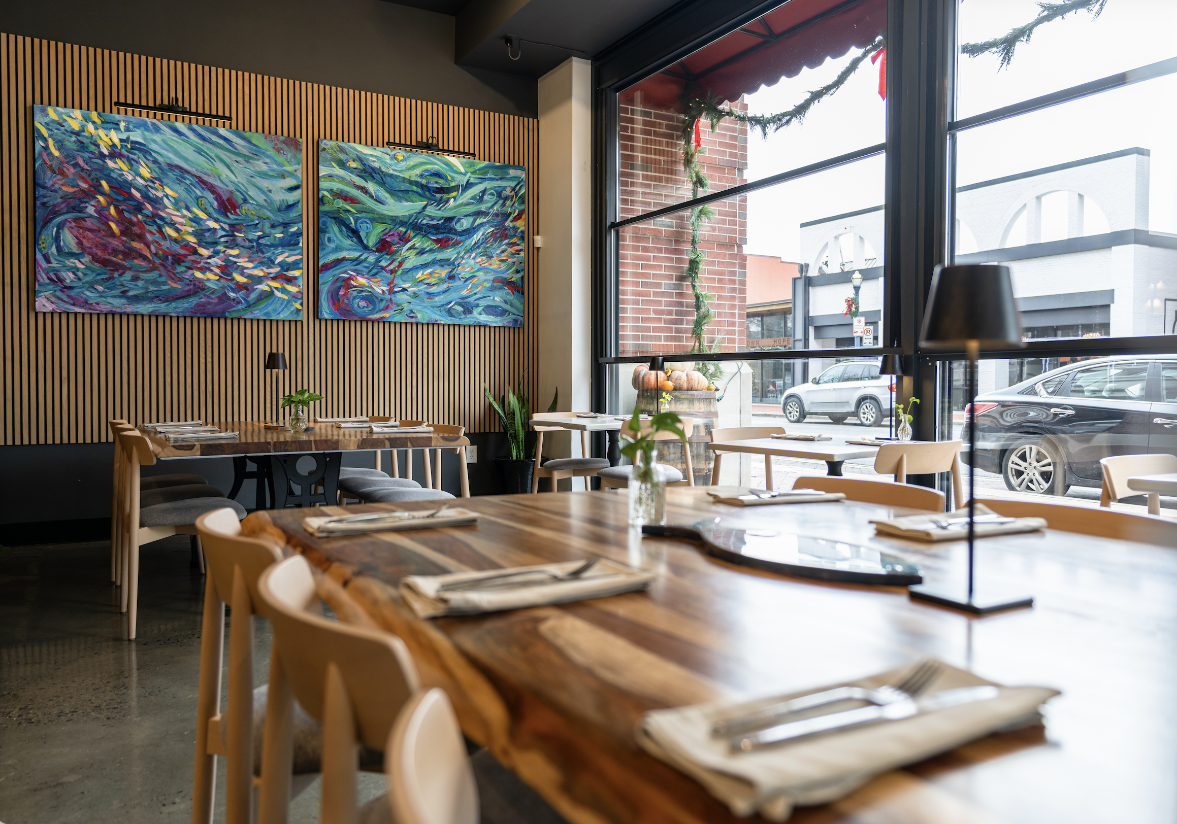 Interior of a restaurant with wooden tables, beige chairs, and large windows decorated for Christmas with garland and pumpkins outside, and abstract blue paintings on the wall.