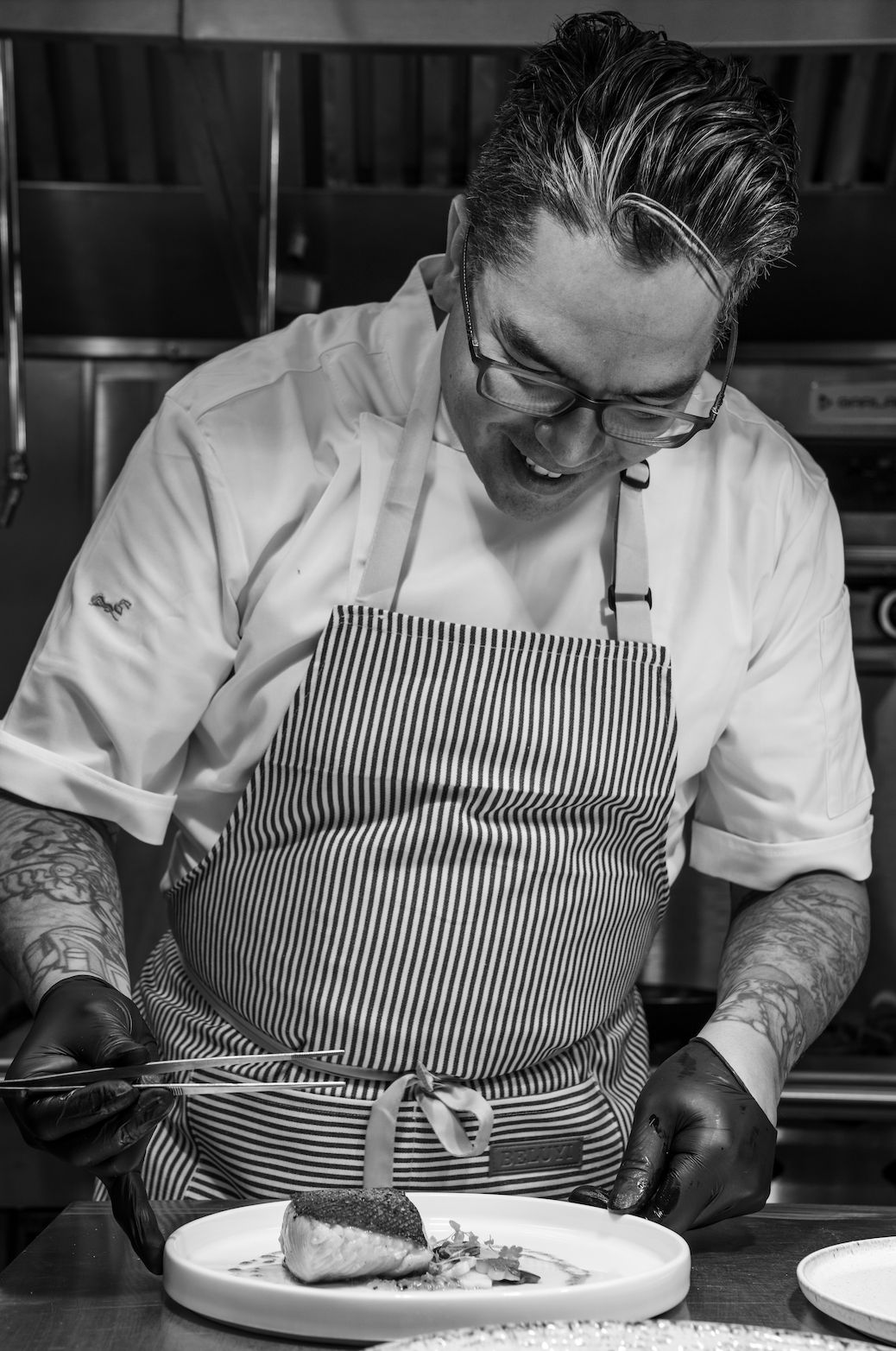 A chef with glasses and tattoos, wearing a striped apron, is garnishing a plated dish with a smile in a professional kitchen.