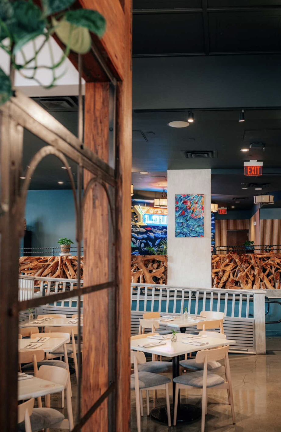 Interior of a restaurant with white tables and chairs, decorative wooden railing, colorful paintings, and neon signs in the background.