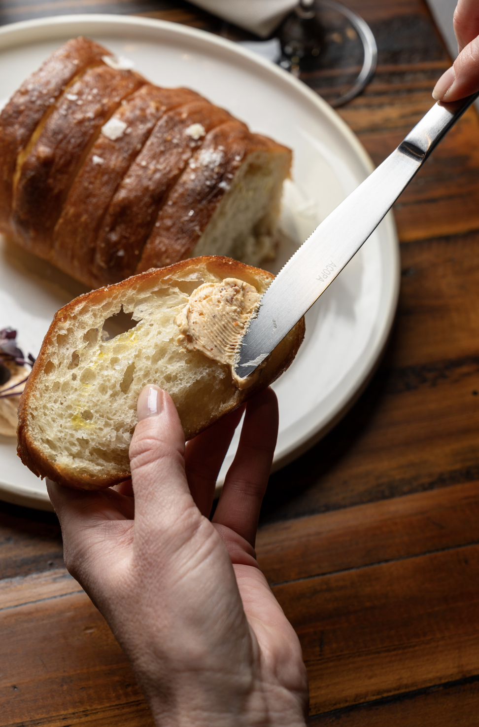 Person spreading butter on a slice of bread with a loaf of bread in the background on a white plate.