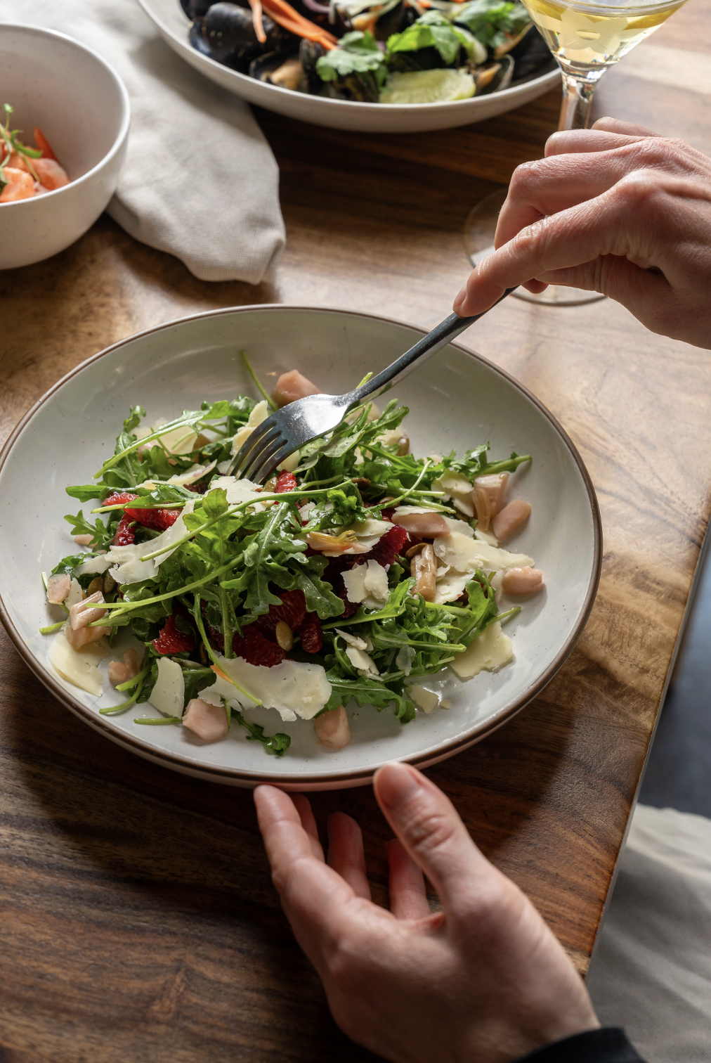 Person eating a salad with greens, cheese, and cranberries, with a glass of white wine nearby.