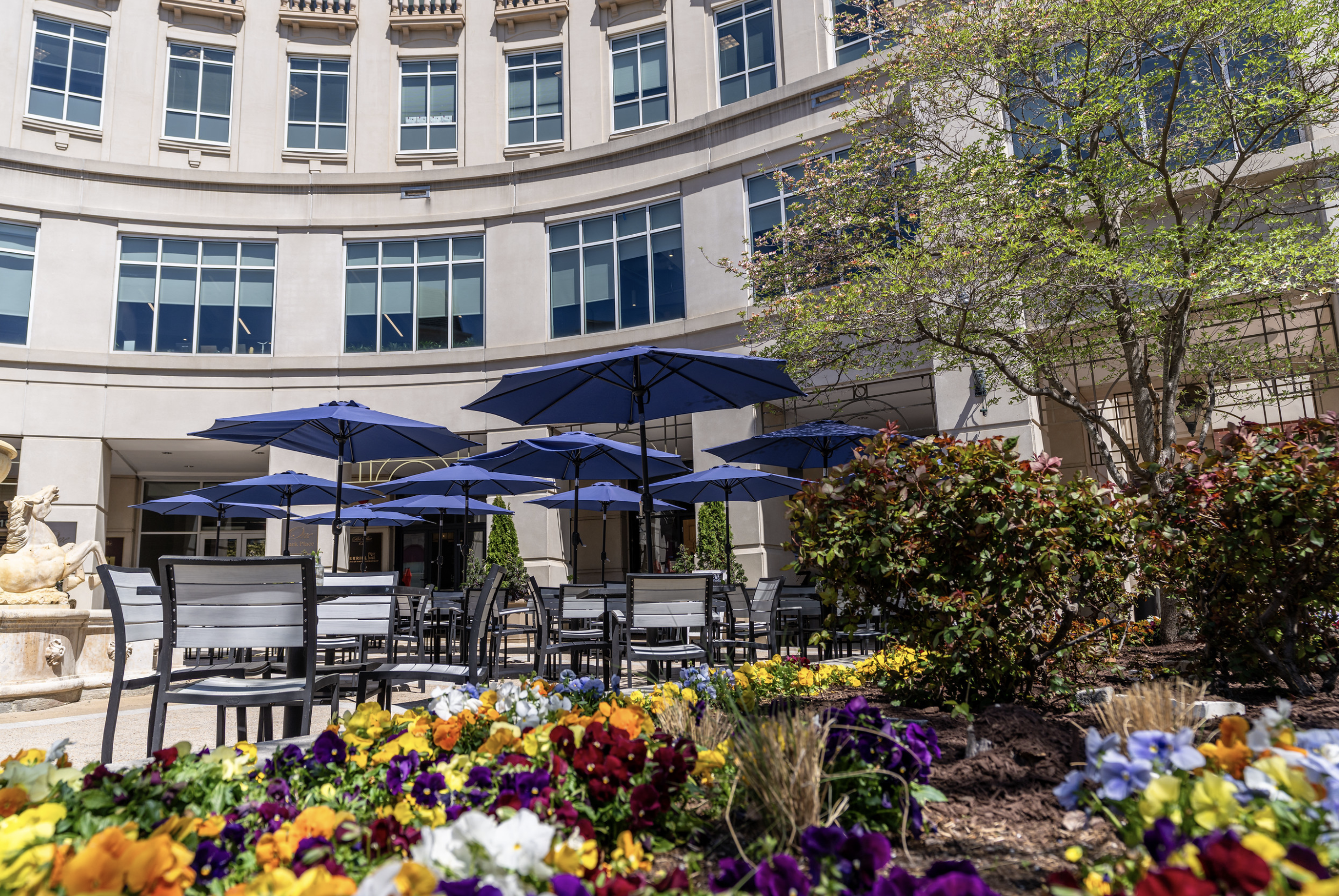 Outdoor cafe seating area with tables, chairs, and large blue umbrellas, surrounded by colorful flowers and green trees, in front of a tall, beige building with multiple windows.