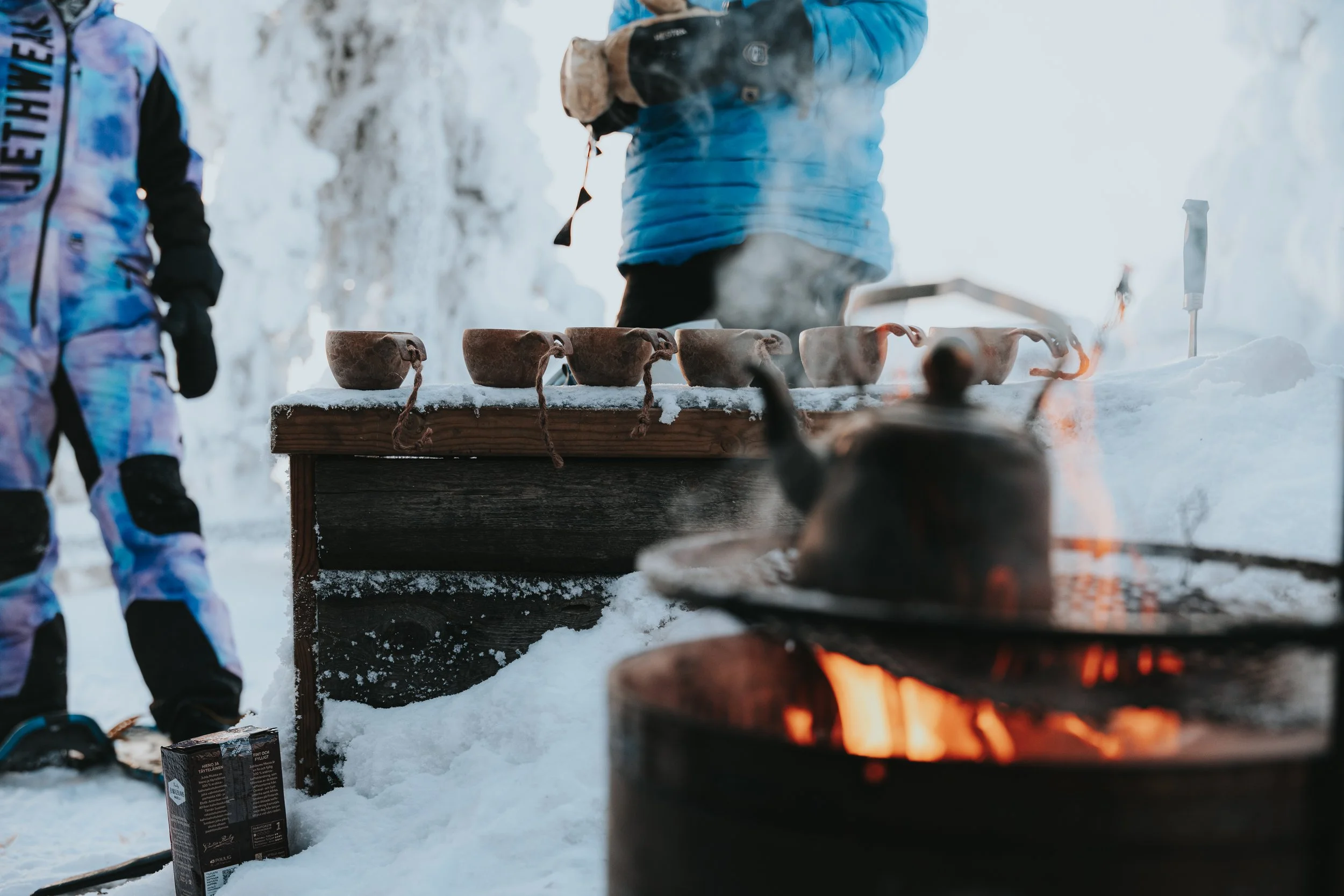 People in winter jackets and gloves standing around a campfire in a snowy outdoor setting, with a table with clay cups and a stove or heater in the background.
