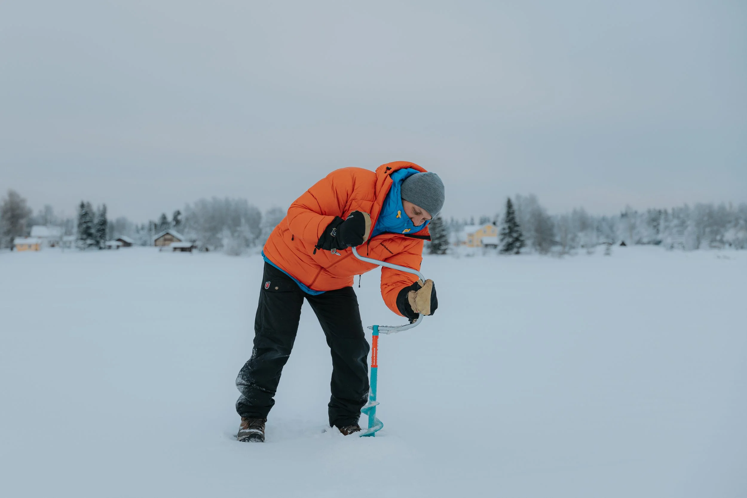 Person in orange jacket and gray beanie using a ice drill to make hole to Tornio river ice. He is going to try to ice fish in Tornio river.