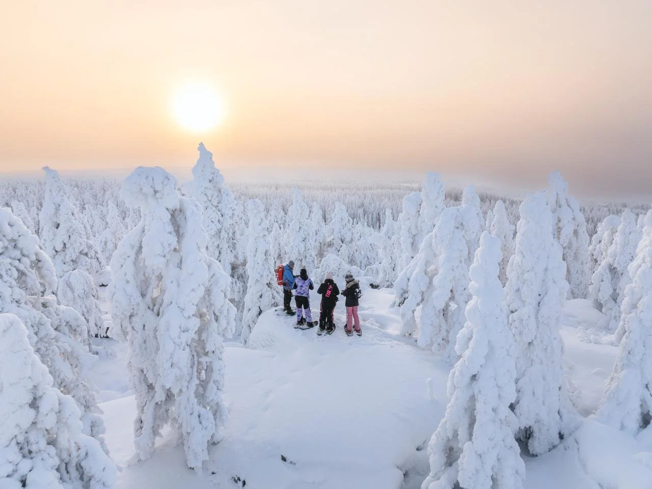 A group of people standing on a snow-covered trail surrounded by snow-laden trees, under a setting sun with a hazy sky.