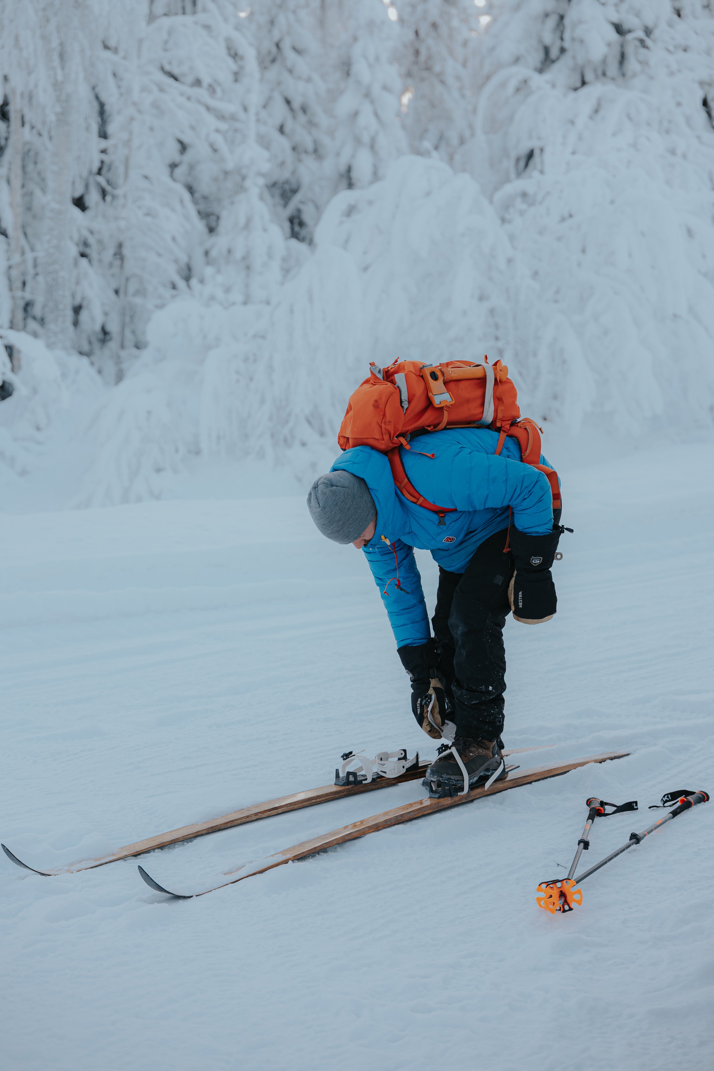 A person dressed in winter gear, including a blue jacket and gray hat, is bending over on a snow-covered landscape, preparing to ski with a backpack on. There are snow-covered trees in the background and ski equipment, including skis, poles, and a he